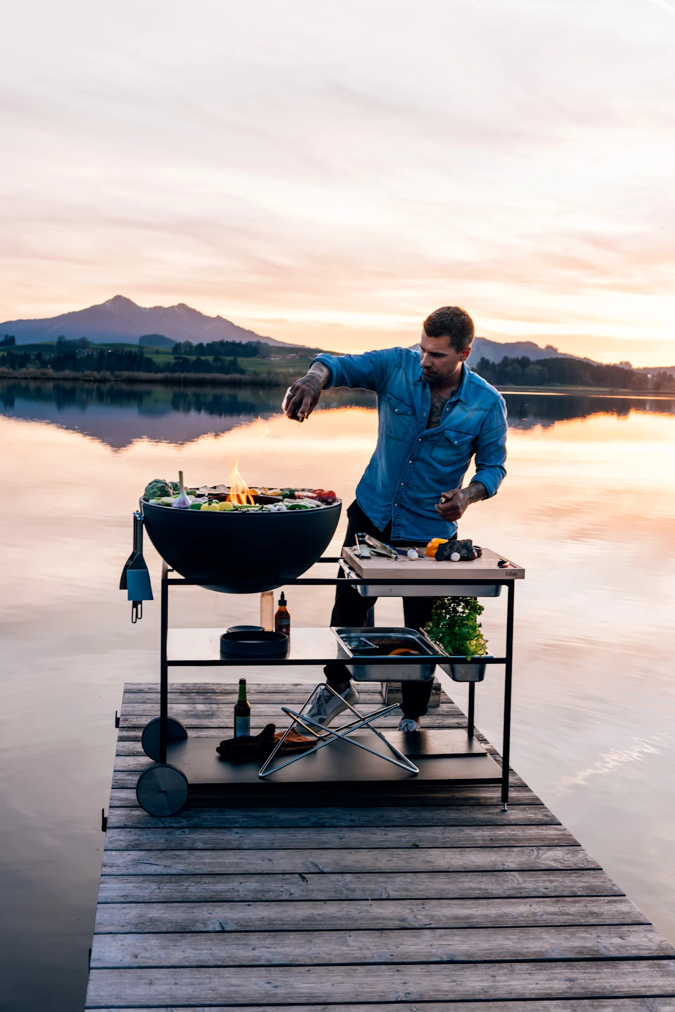 Hombre con camisa vaquera asando comida en una parrilla redonda negra y mesa de preparación en un muelle de madera al atardecer.