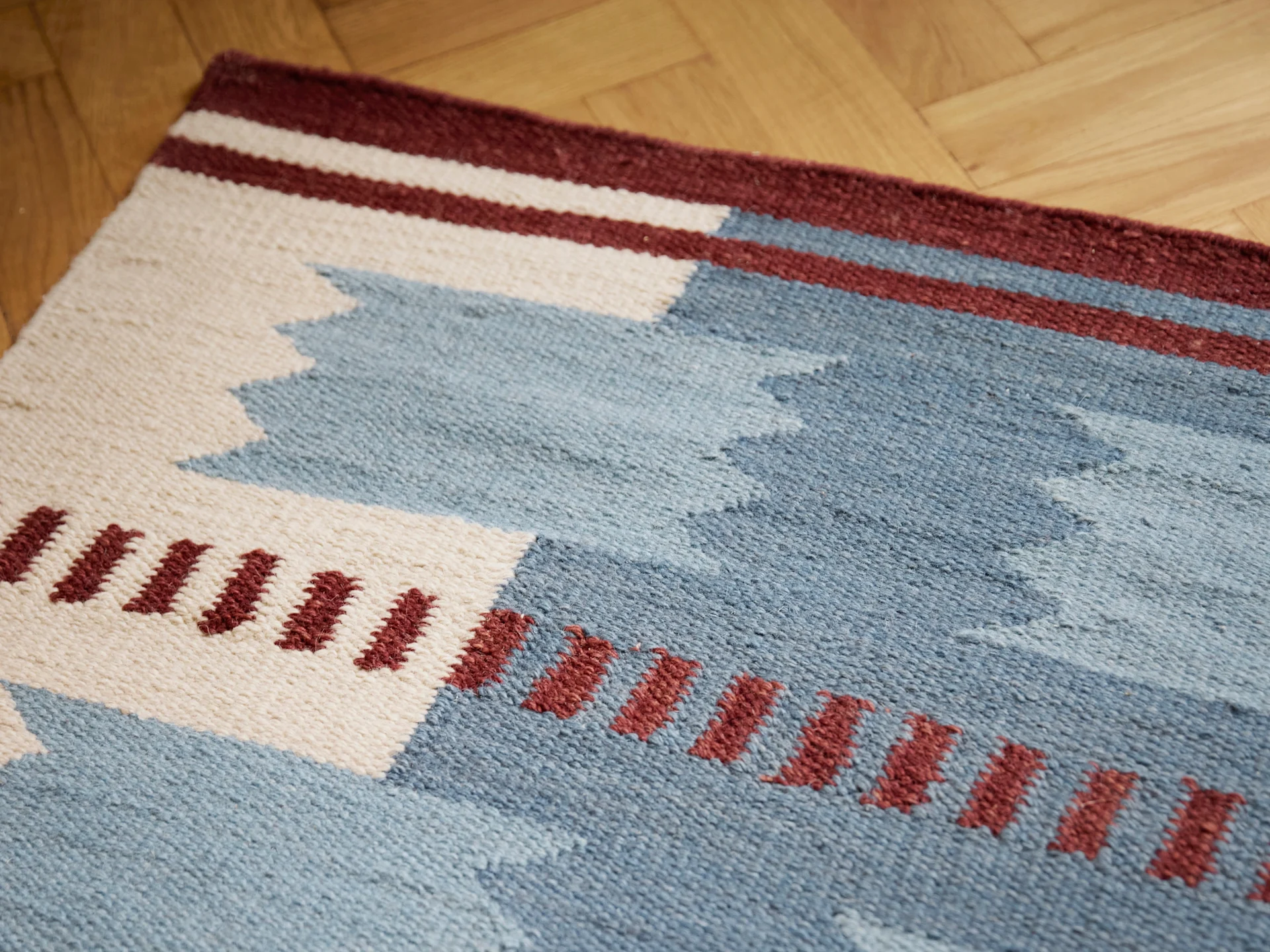 Alfombra tejida con patrones geométricos en azul, blanco y burdeos sobre un suelo de madera.