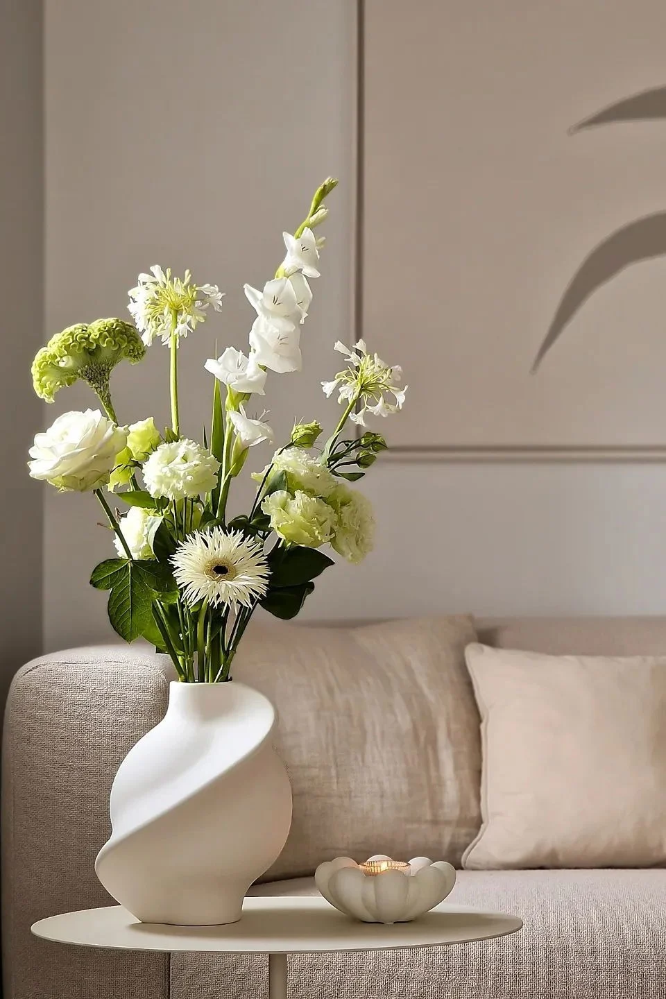 En un salón de color beige, una mesa auxiliar está decorada con un jarrón de flores cortadas y un portavelas para tealight.