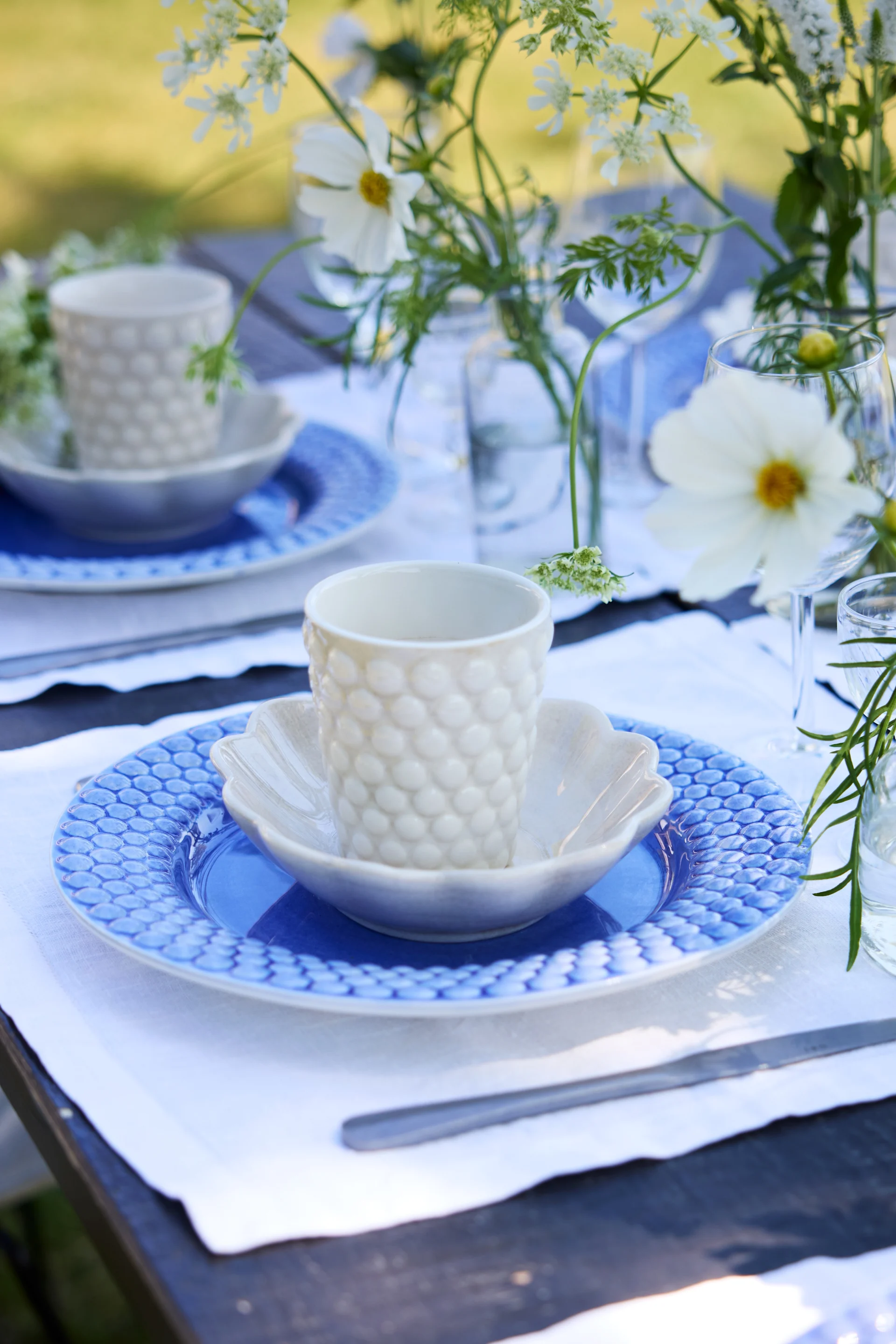 Primer plano de una mesa de exterior en azul y blanco. Un plato Bubbles azul junto con una taza Bubbles blanca sobre una mesa decorada con flores.
