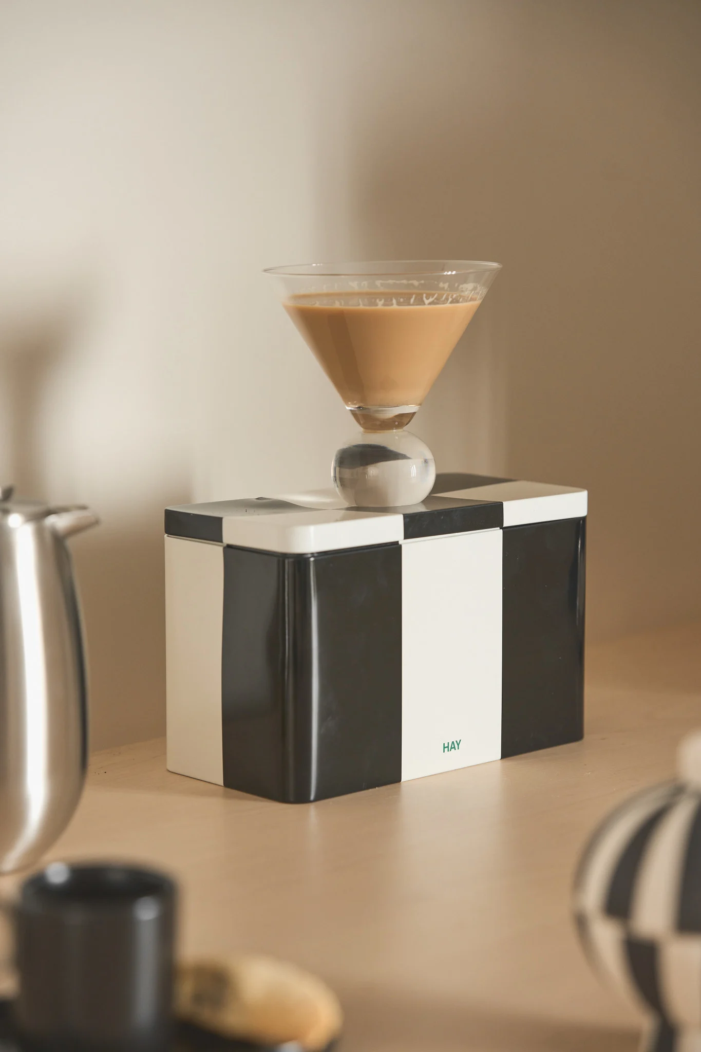 Copa de martini con bebida marrón claro y tallo esférico sobre una caja a cuadros blancos y negros con logo 'HAY'.