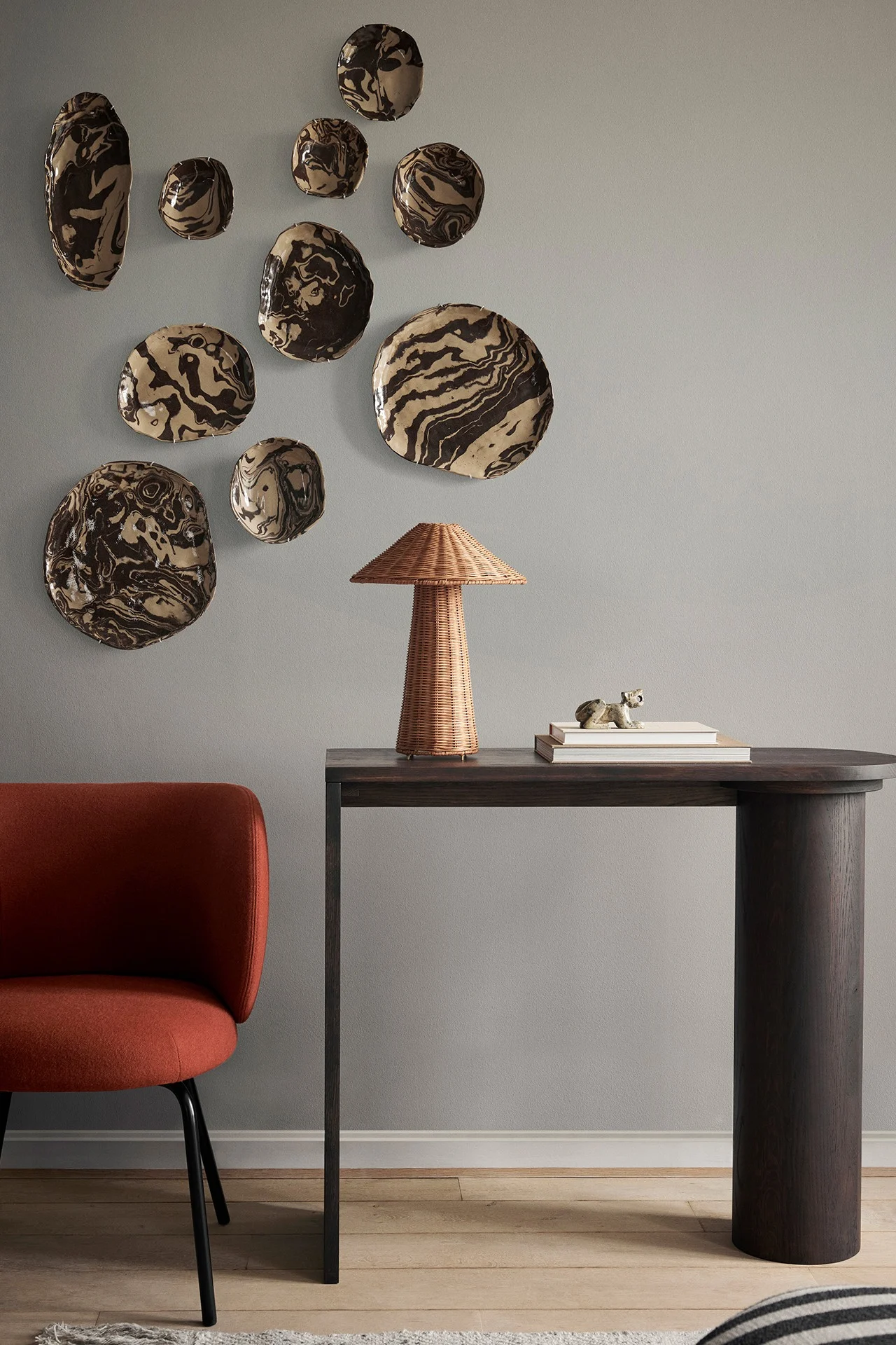 Pared gris claro con platos de cerámica marmoleados, una mesa consola de madera oscura con lámpara de mimbre y una silla color óxido.