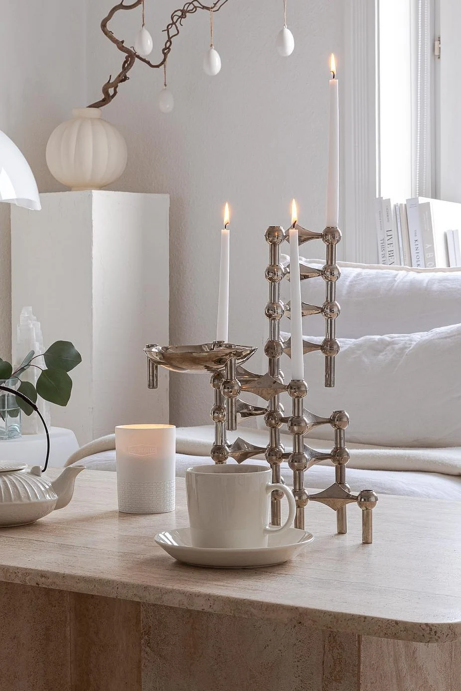 Candelabro modular plateado moderno con tres velas blancas encendidas, taza de té y tetera sobre una mesa de piedra con decoración de Pascua.