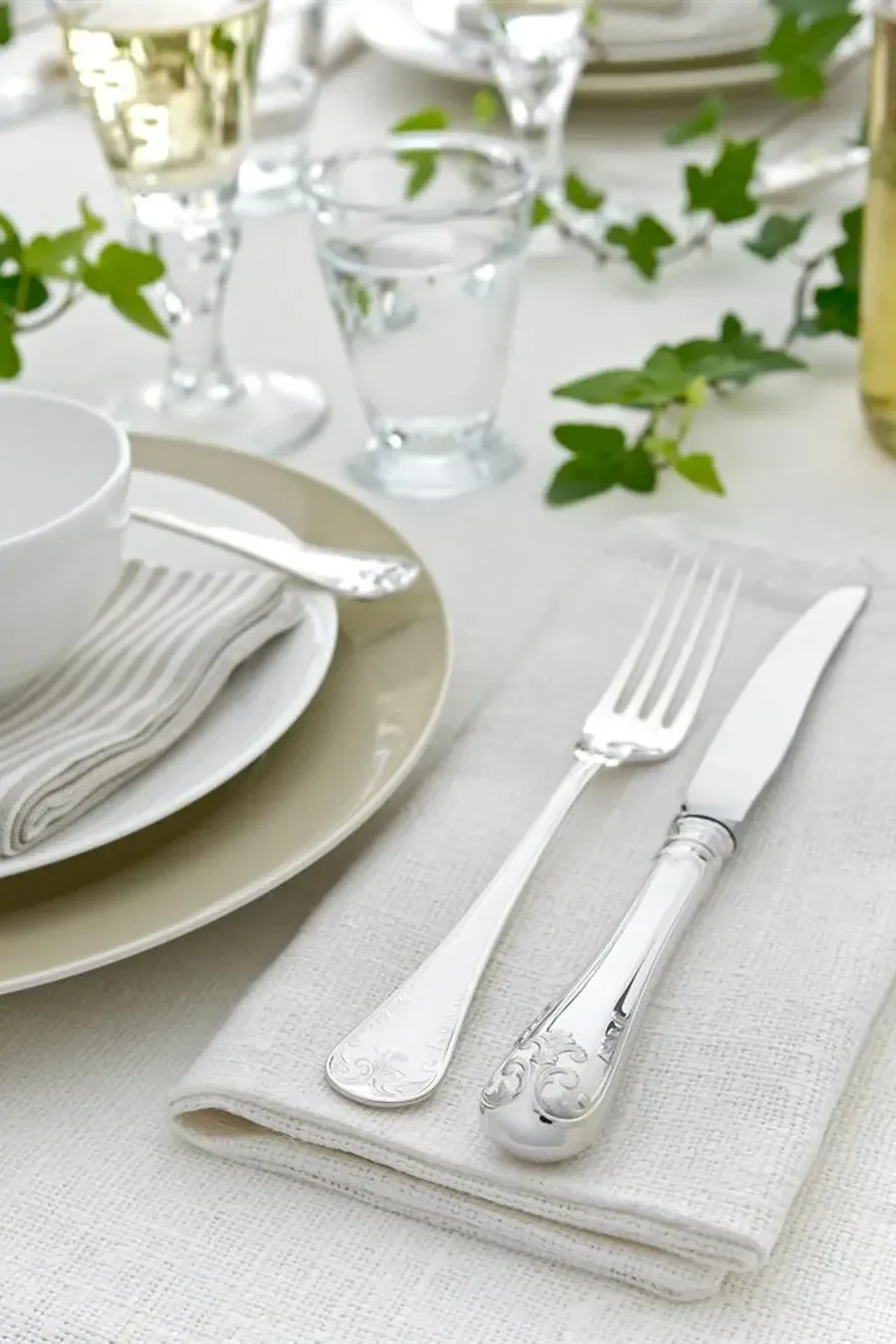 Elegante mesa puesta con cubiertos de plata ornamentados, platos apilados, copas y hiedra verde sobre un mantel blanco.