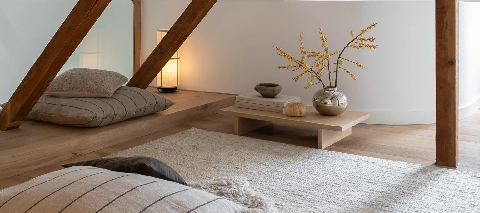 Una habitación acogedora con suelos de madera clara y paredes blancas, con cojines de suelo, una lámpara encendida y una mesa baja con un jarrón plateado con bayas amarillas.