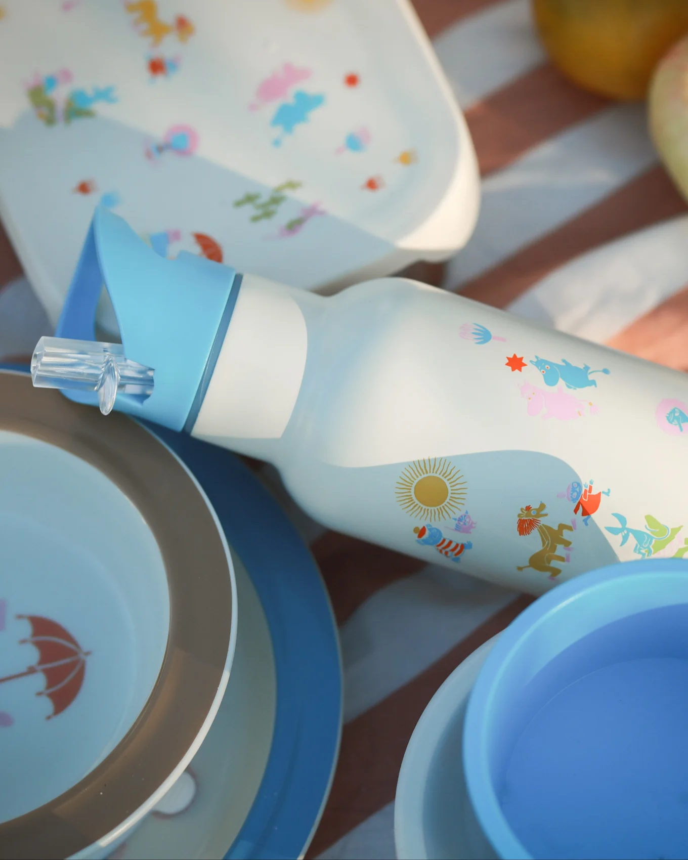 Botella de agua blanca con pajita azul, personajes de dibujos animados coloridos y sol, rodeada de platos de picnic a juego sobre una manta a rayas.