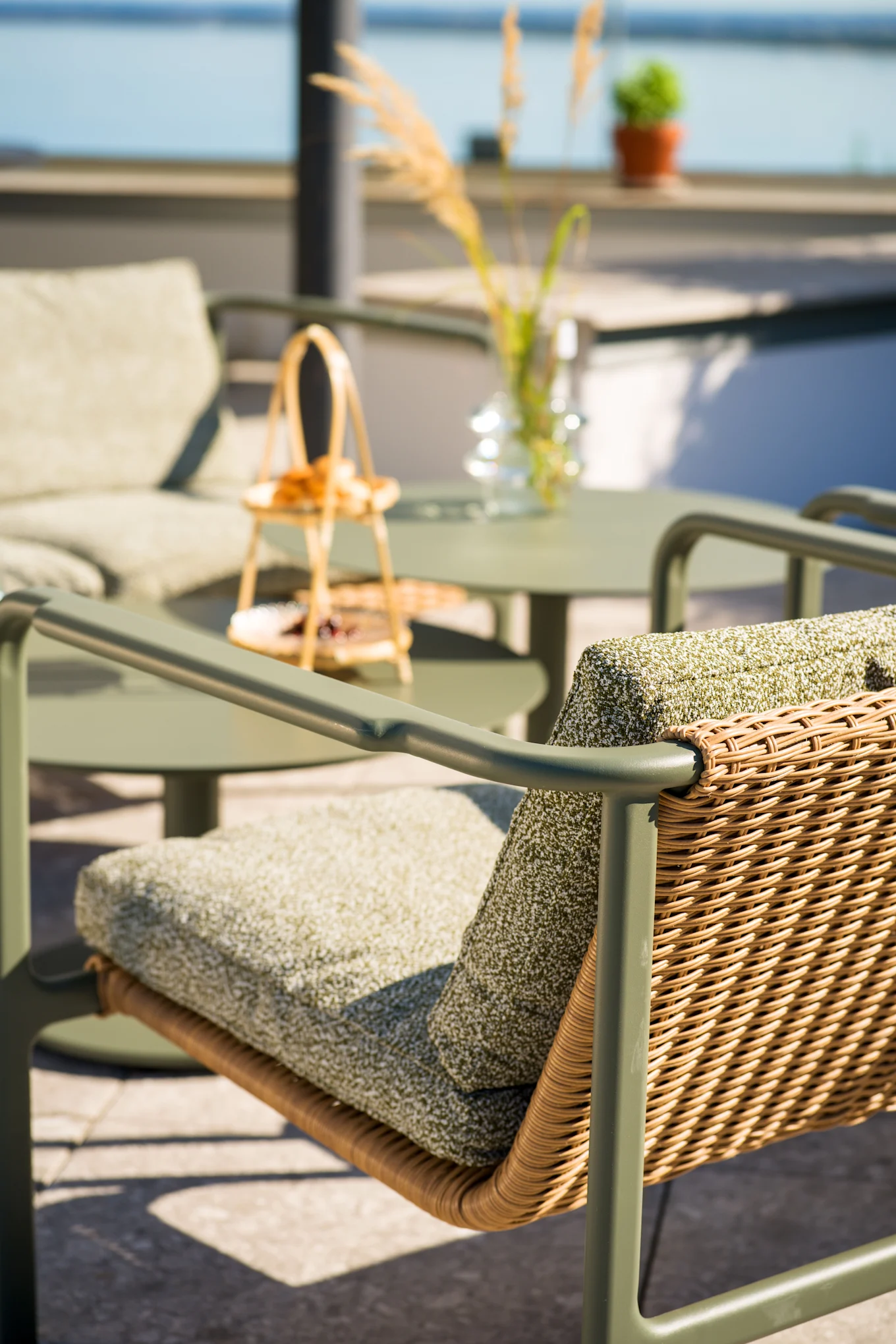 Silla de patio exterior con estructura verde oliva, respaldo de ratán trenzado y cojines verdes. Mesa y silla a juego en fondo borroso cerca del agua.