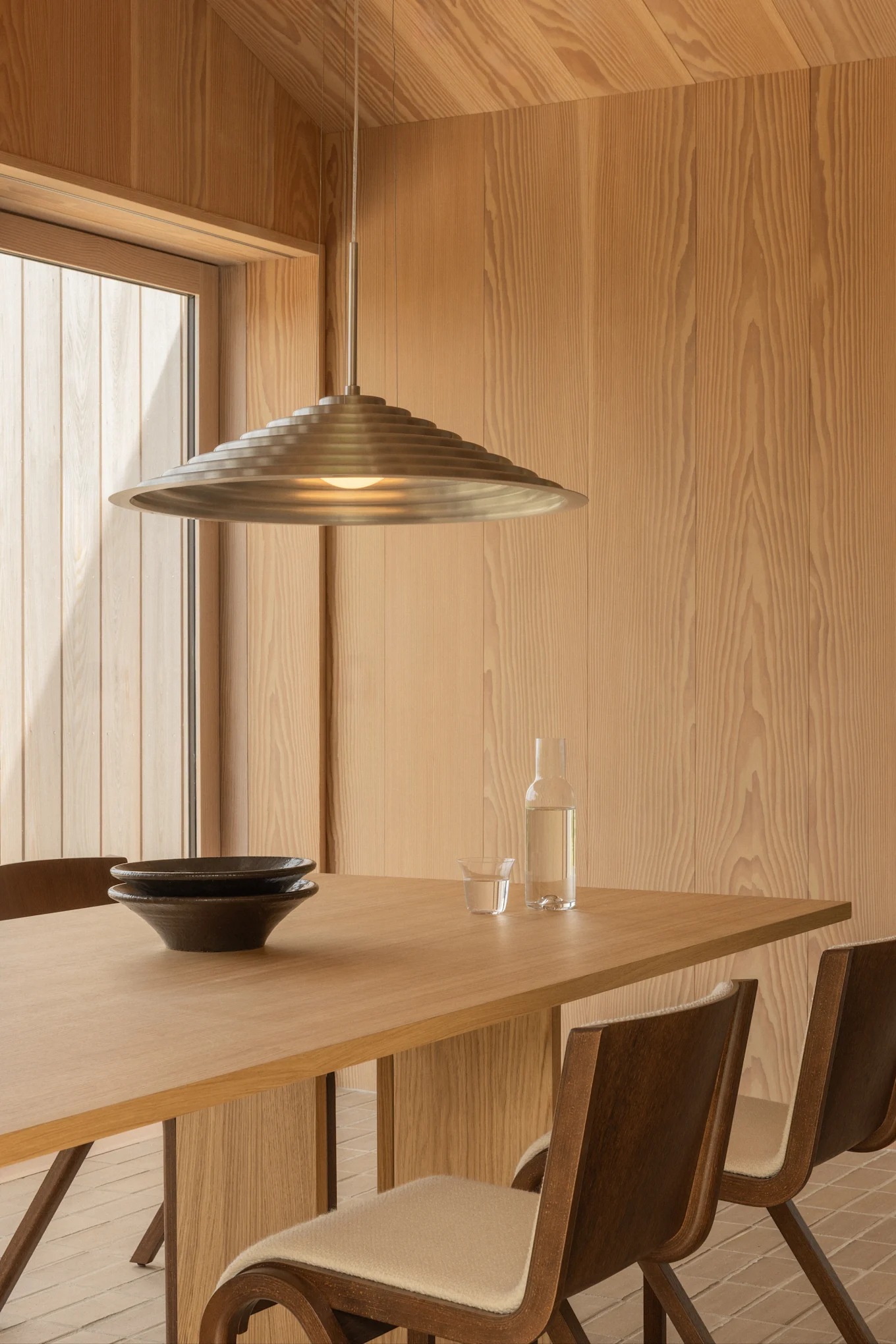 Comedor moderno con paredes de madera clara, una mesa de madera con dos cuencos oscuros, una botella de agua y una lámpara colgante de metal escalonada.