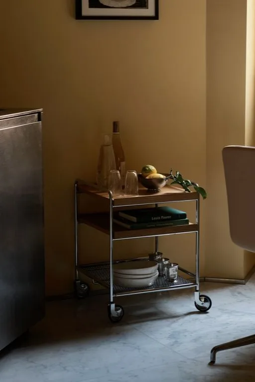 Un carrito auxiliar de tres niveles con estructura de metal y estantes de madera, que contiene botellas, vasos, limones, libros y platos.