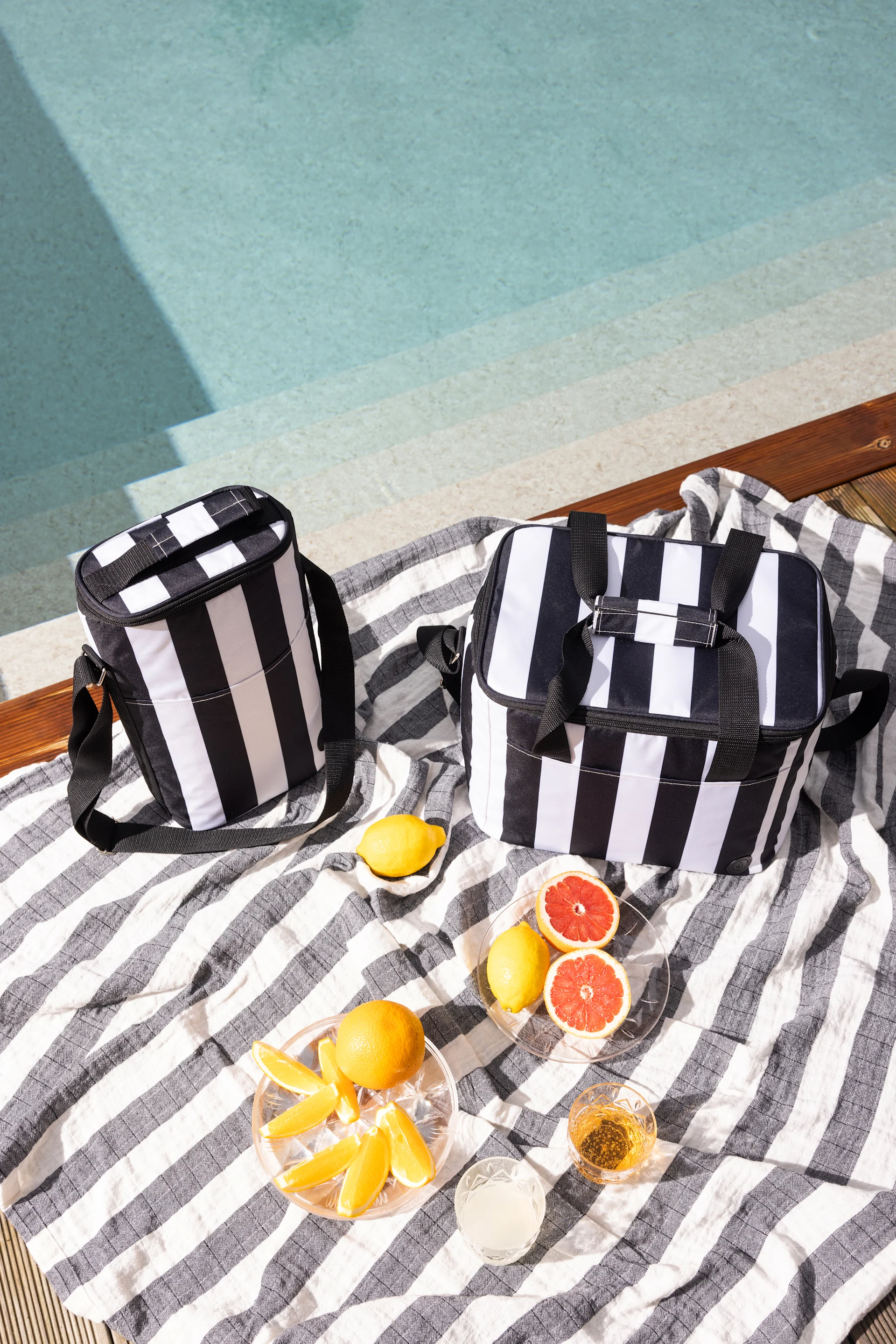 Dos bolsas térmicas a rayas blancas y negras sobre una toalla a rayas junto a una piscina, con cítricos y bebidas.