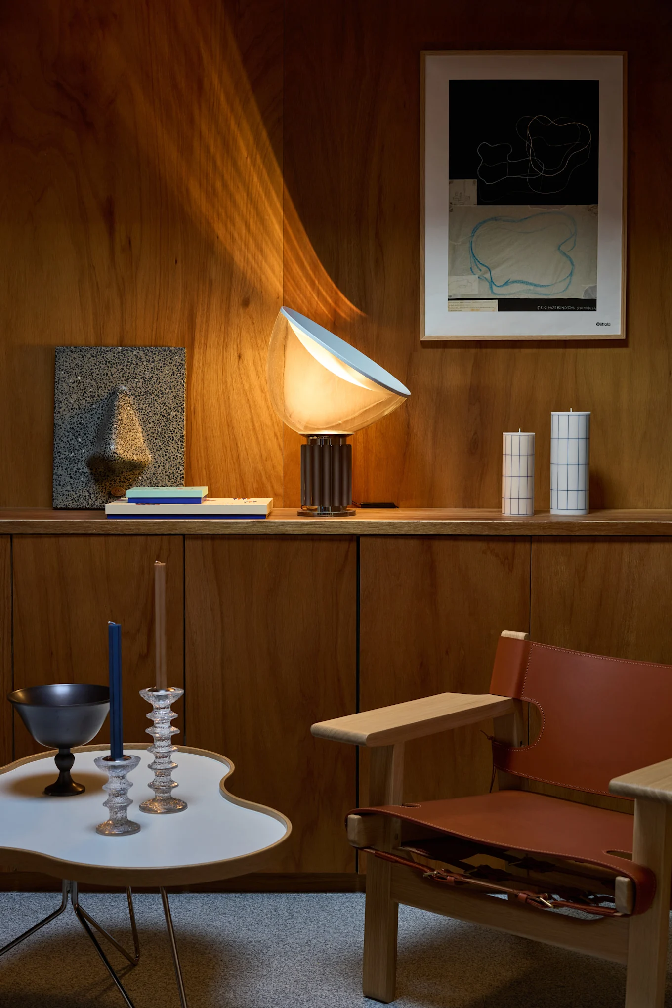 Un salón con paredes de madera, una lámpara de mesa brillante, arte abstracto de pared, velas decorativas, una mesa auxiliar moderna y un sillón de cuero.