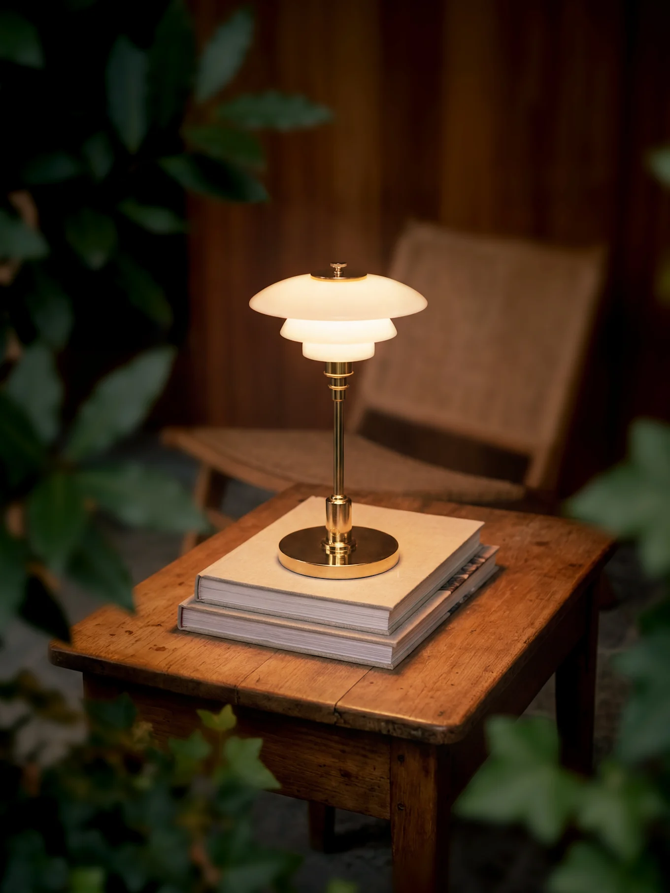 Una lámpara de mesa de latón brillante de varios niveles se asienta sobre dos libros en una mesa de madera, rodeada de follaje oscuro.