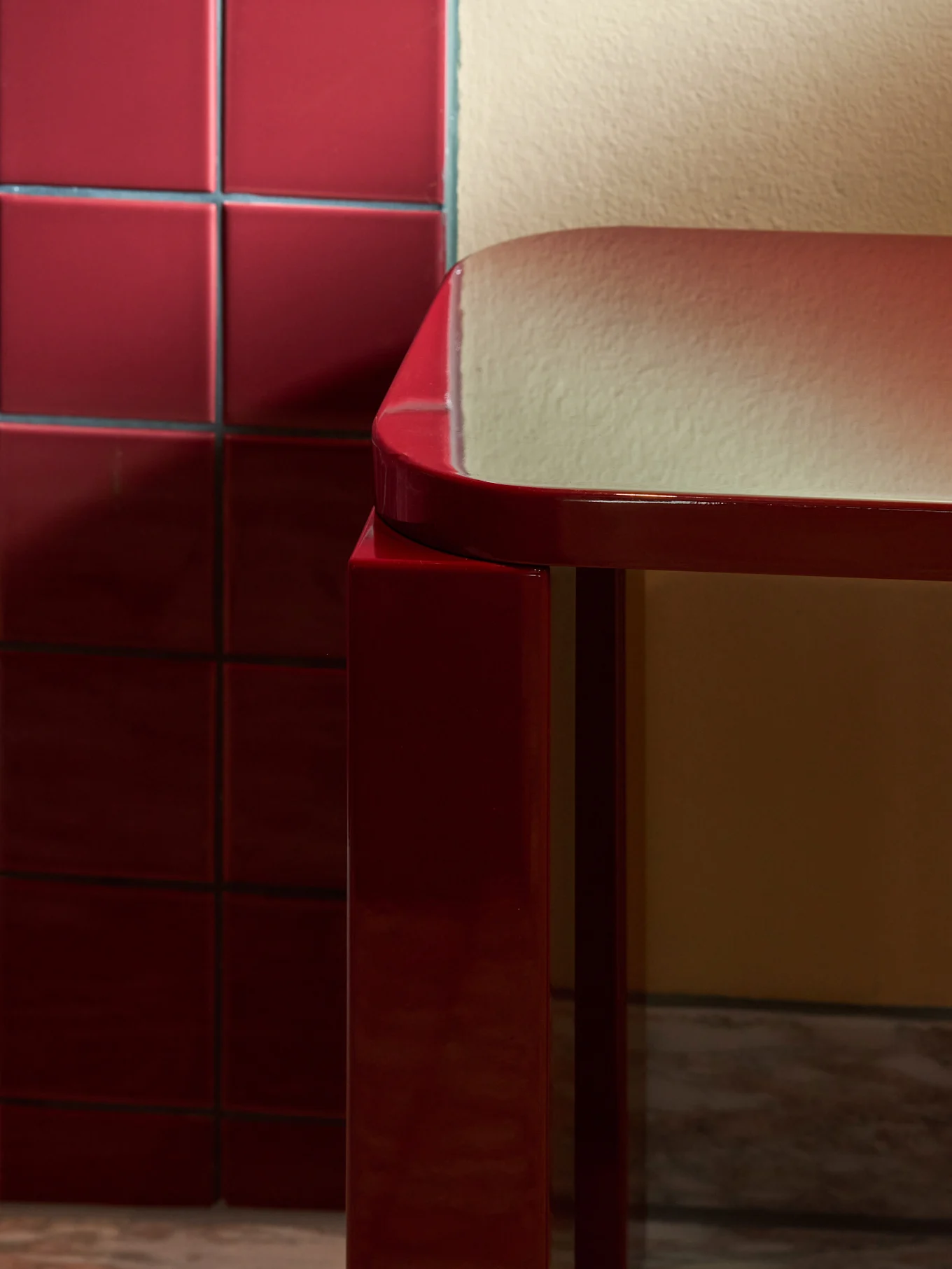 Elegante y brillante taburete rectangular rojo con bordes redondeados sobre suelo de mármol, junto a una pared de azulejos rojos.