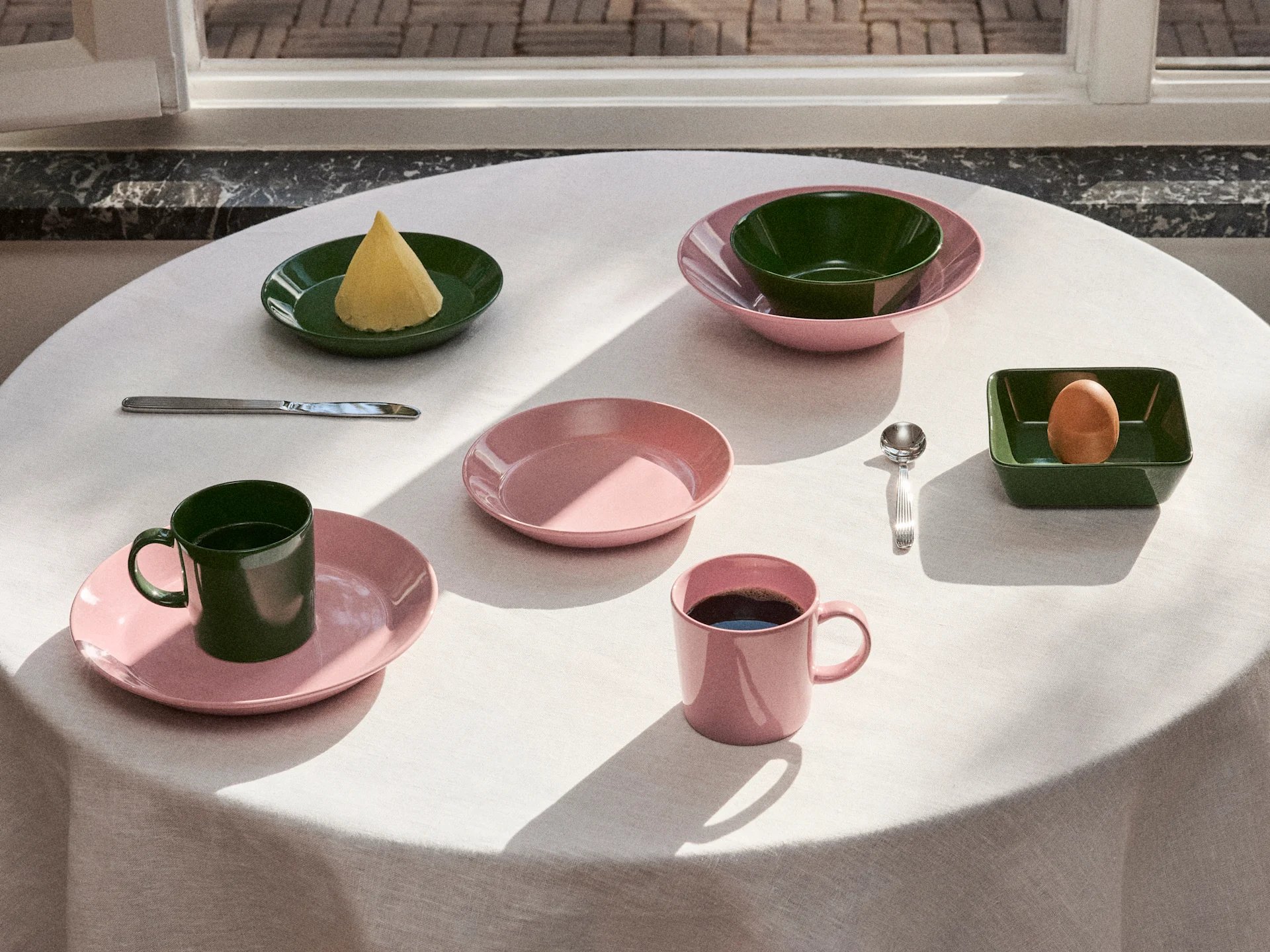 Mesa puesta con vajilla rosa y verde oscuro, que incluye platos, cuencos y tazas, con mantequilla, un huevo y café bajo luz natural.