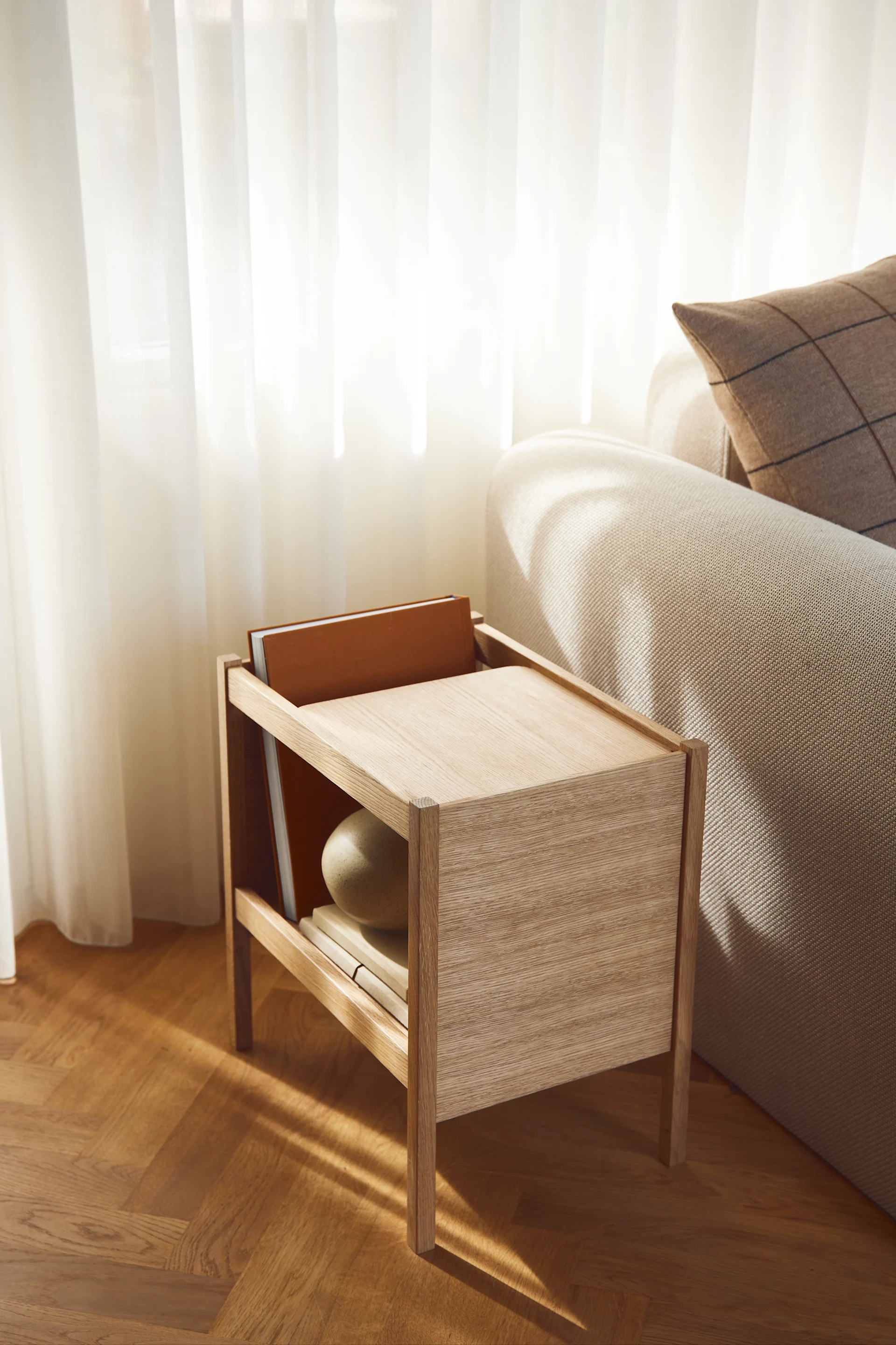 Una mesa auxiliar de madera clara con libros y una esfera decorativa junto a un sofá beige, bañada por la luz del sol de cortinas transparentes.