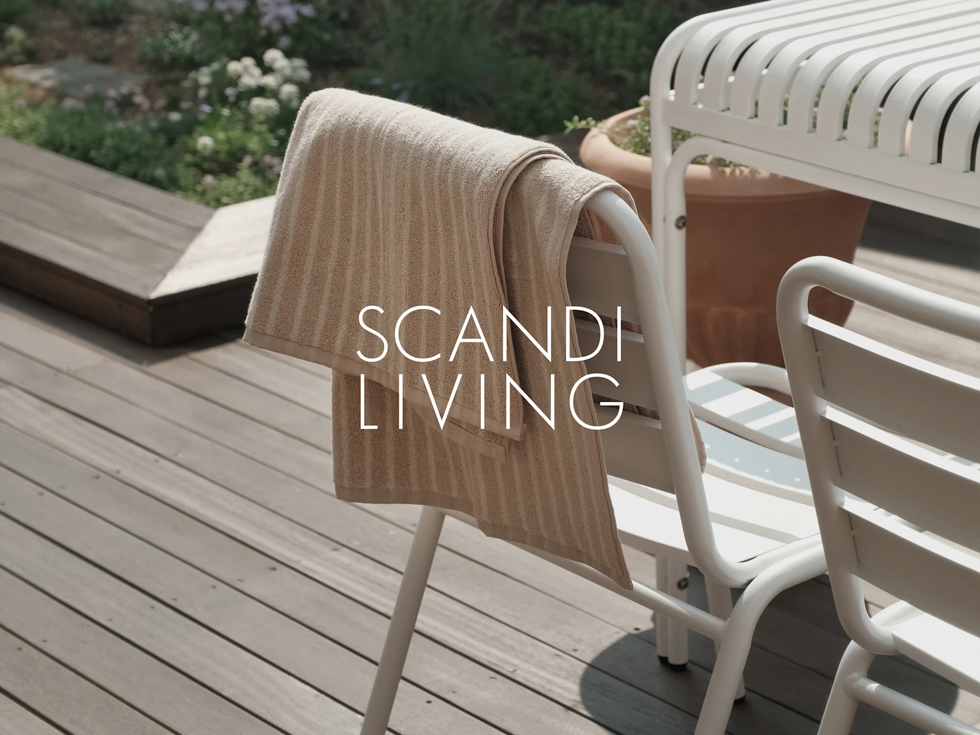 Una toalla a rayas beige cuelga de una silla de exterior blanca en una terraza de madera con plantas. Texto: SCANDI LIVING.