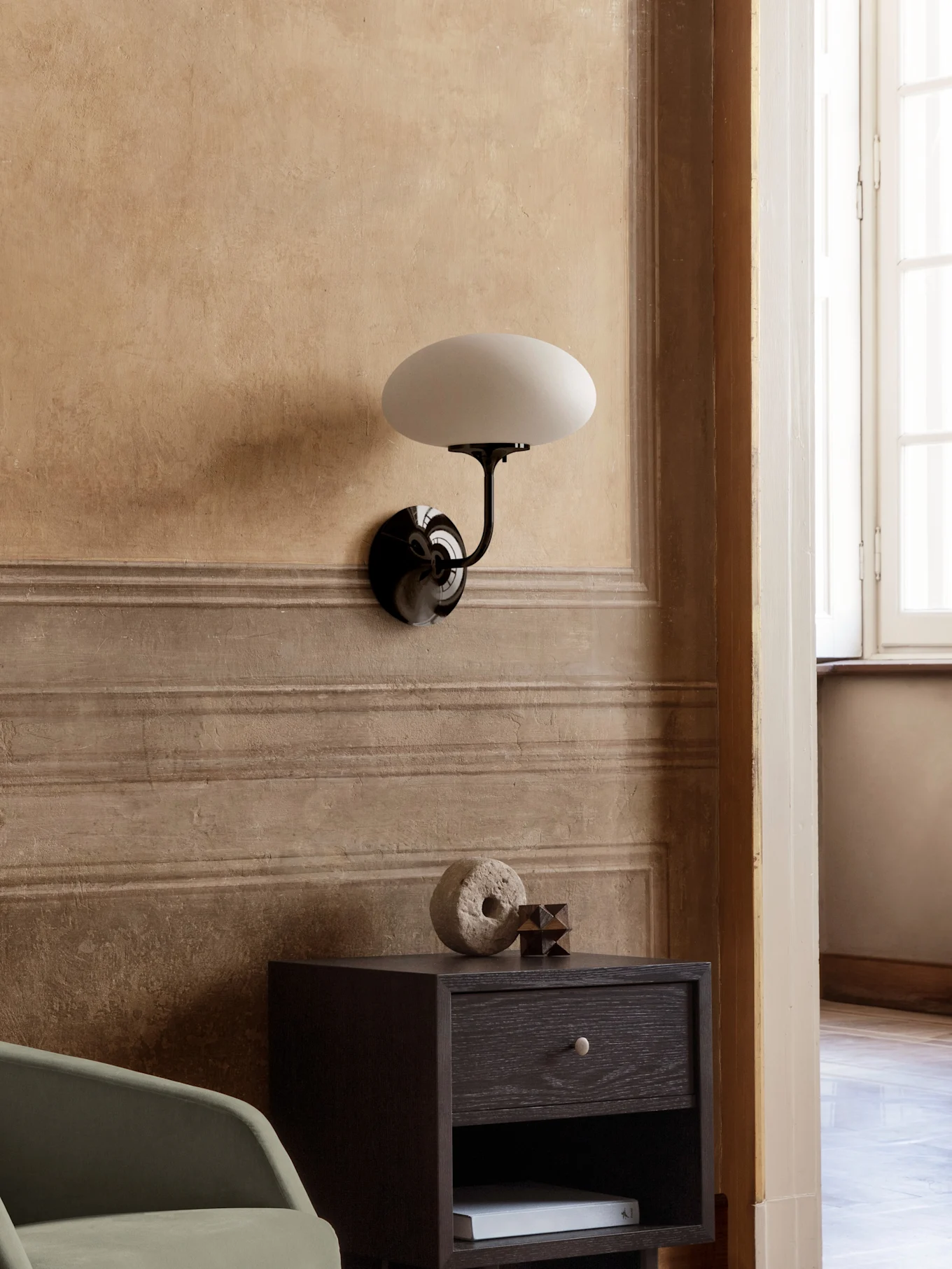 Lámpara de pared con brazo negro y pantalla ovalada blanca sobre una mesita de noche de madera oscura con decoración.
