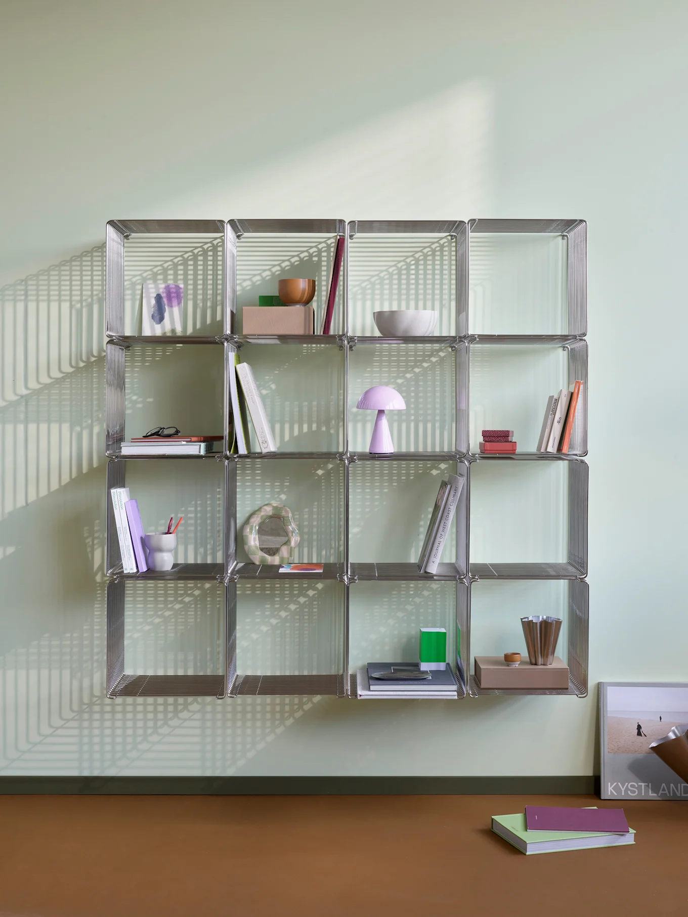 Una estantería modular cromada con libros y objetos decorativos, iluminada por fuertes sombras lineales en una pared verde claro y suelo marrón.