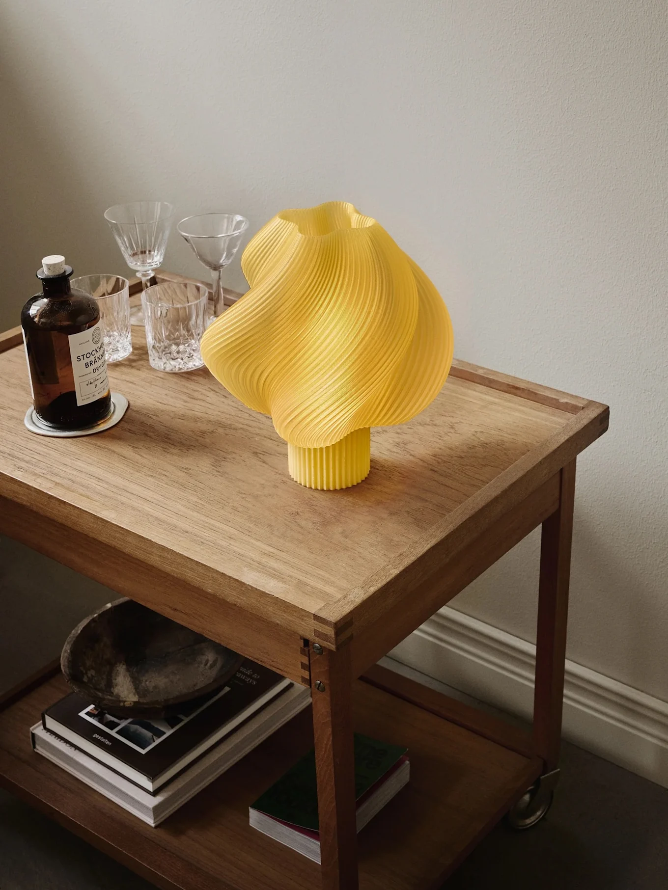 Lámpara amarilla plisada sobre carrito de madera con botella de ginebra, vasos y libros en el estante inferior.