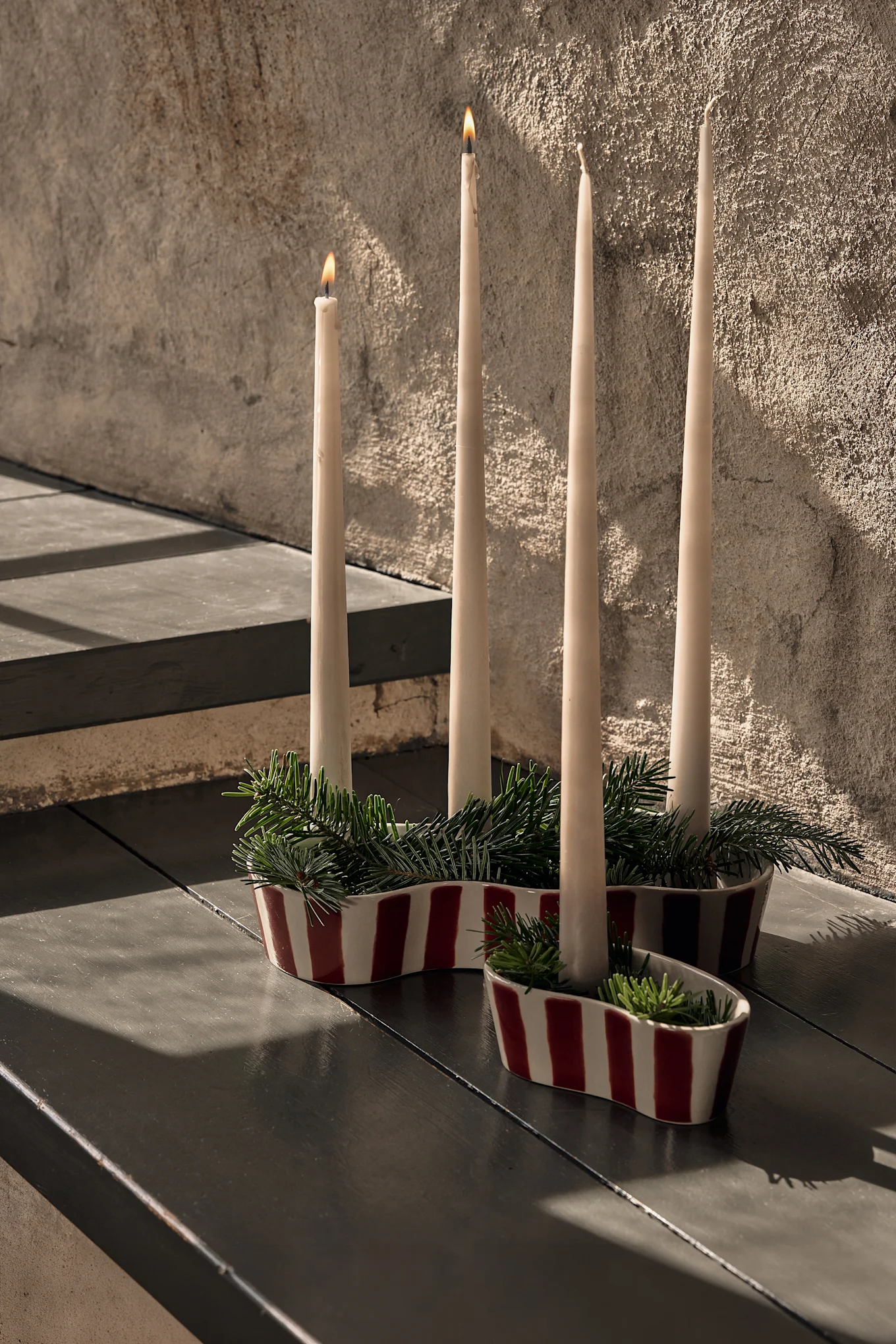 Cuatro velas crema en portavelas de cerámica a rayas rojas y blancas con ramas de pino, dos velas encendidas, sobre una mesa oscura.