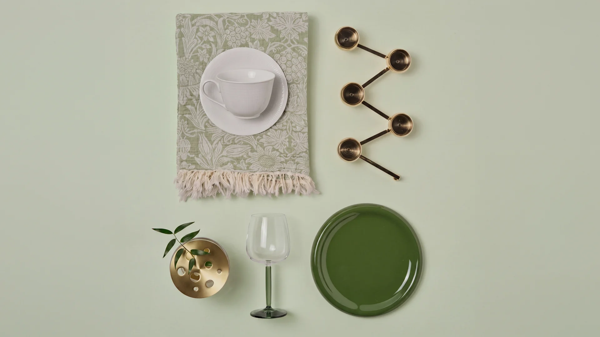 Taza blanca, servilleta verde, cucharas latón, jarrón con planta, copa verde, plato verde oscuro.