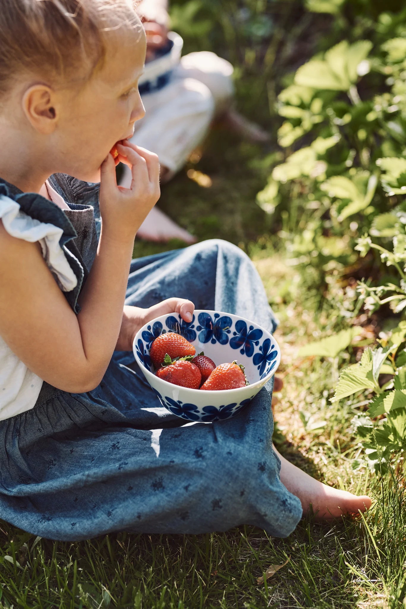 Suecia también ofrece muchas fresas en verano. Aquí, una niña está sentada en un prado, sosteniendo en la mano el cuenco Mon Amie de Rörstrand, que está lleno de fresas.