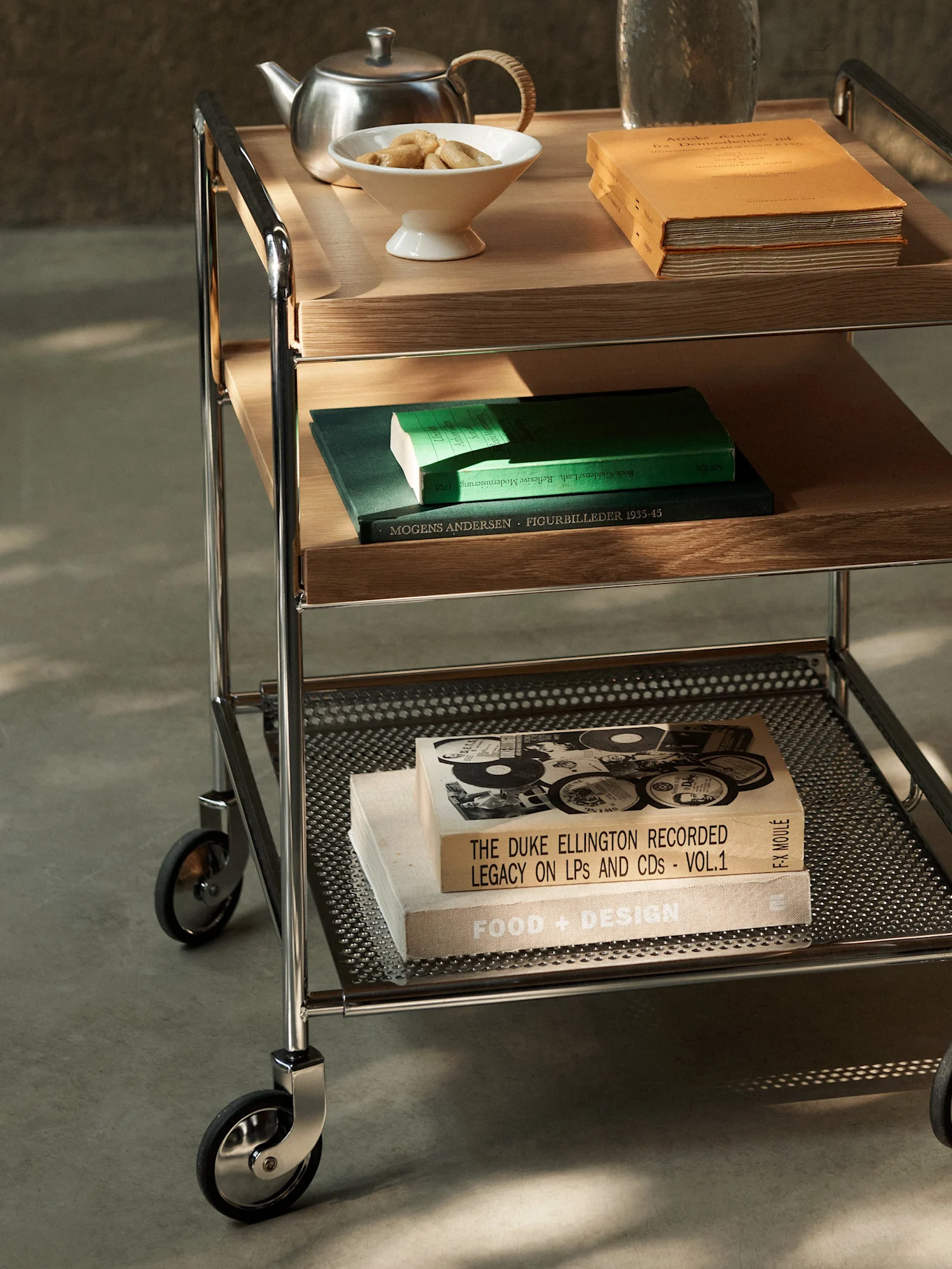 Un carrito de servir de tres niveles con estructura cromada, estantes de madera y metal perforado, con libros, tetera y galletas.