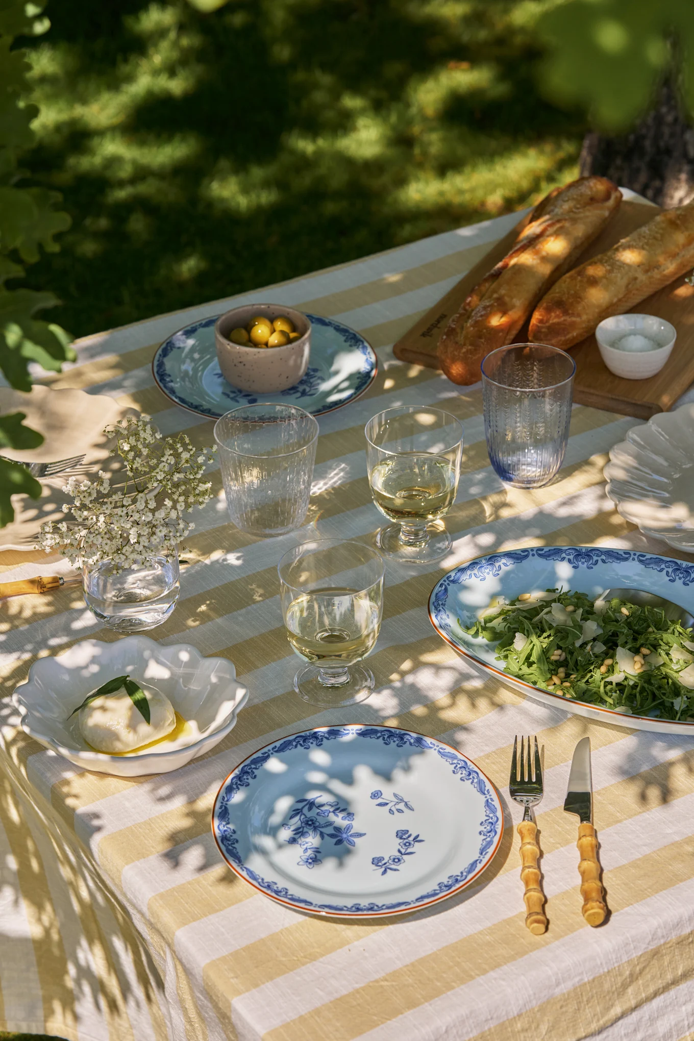 Mesa de comedor al aire libre con mantel de rayas blancas y amarillas, platos azules florales, vino, ensalada, mozzarella, aceitunas y baguettes.