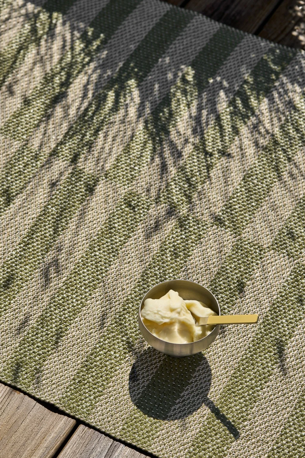 Un cuenco plateado con helado amarillo pálido y una cuchara dorada descansa sobre una alfombra de rayas verdes y beige con sombras de hojas, junto a tablas de madera.