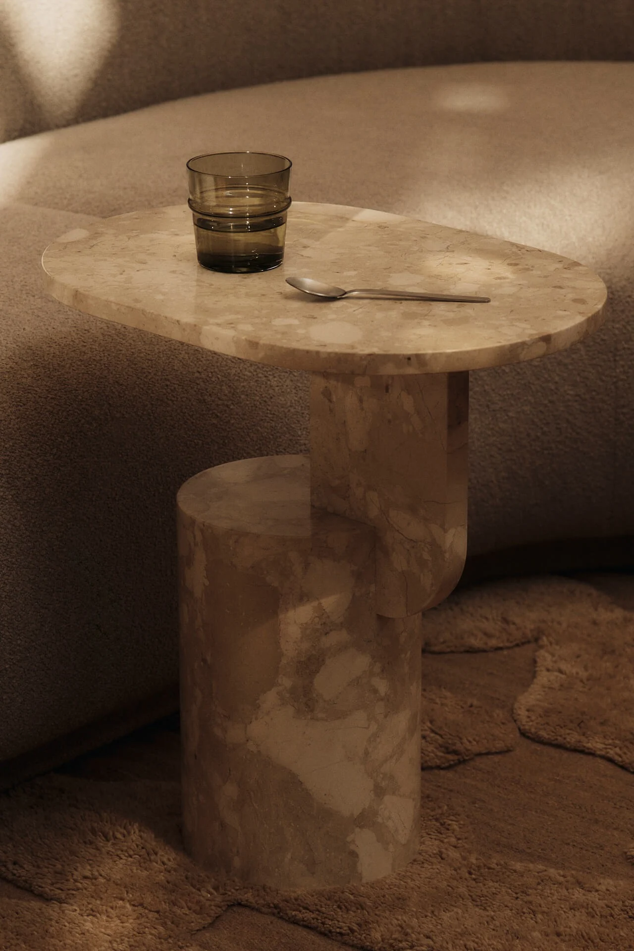 Mesa auxiliar de mármol beige con un vaso de agua verde oscuro y una cuchara plateada, junto a un sofá texturizado y una alfombra.