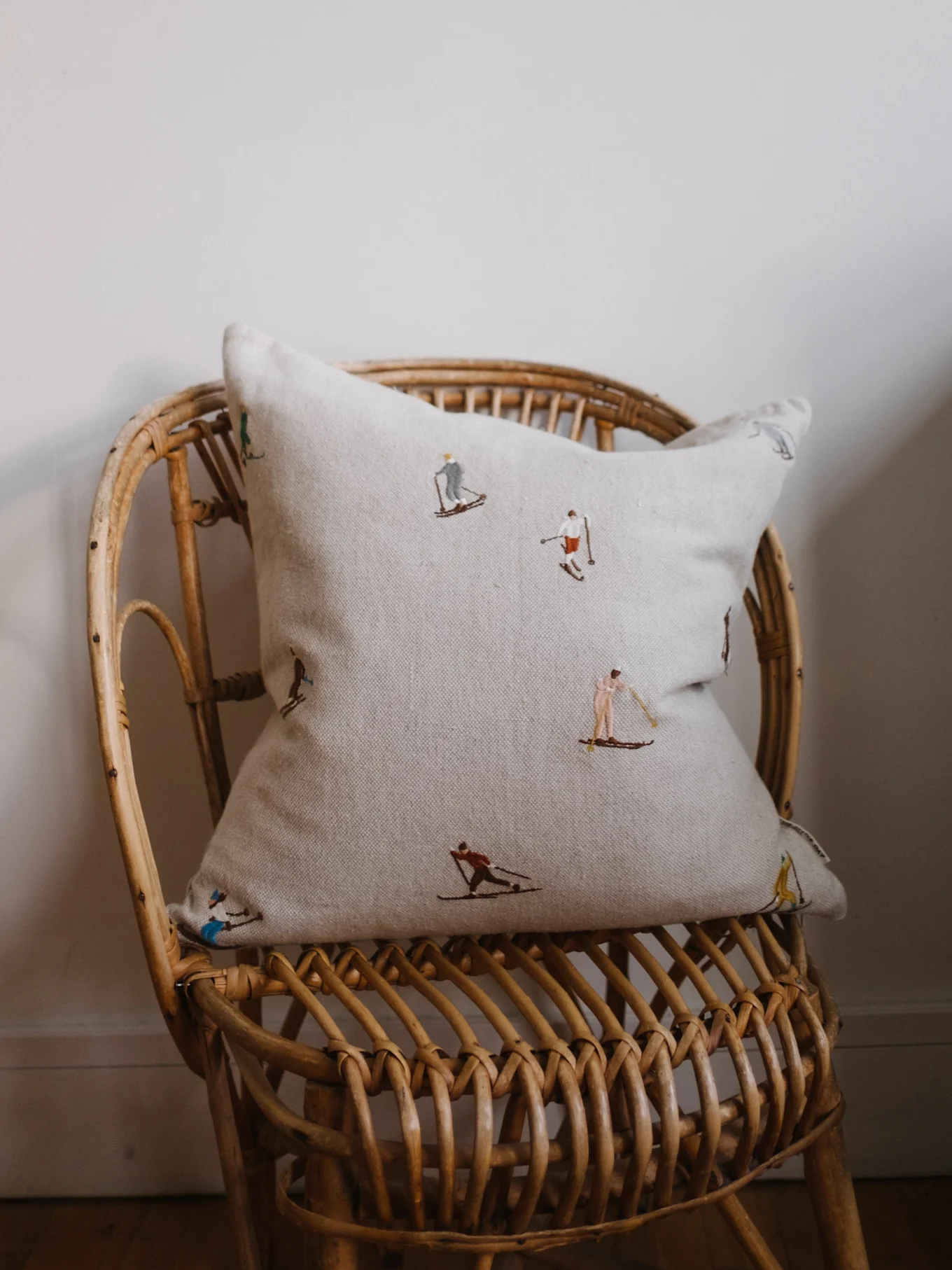 Cojín beige claro con figuras de esquí bordadas en una silla de mimbre frente a una pared blanquecina.