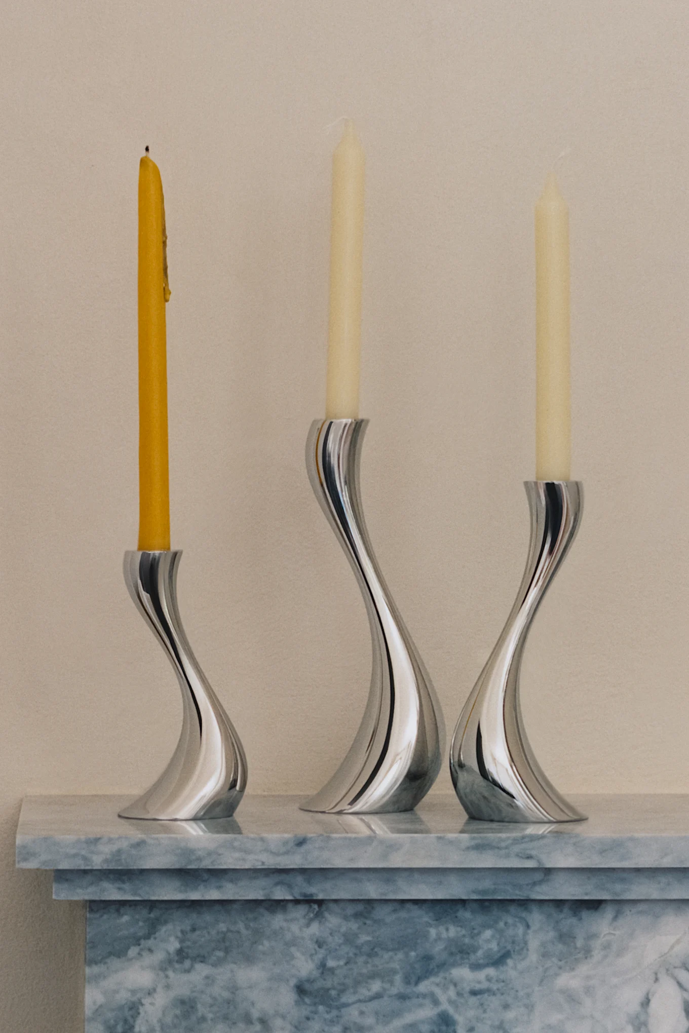 Tres elegantes candelabros plateados y curvos con una vela amarilla quemada y dos velas crema sin encender sobre una superficie de mármol.