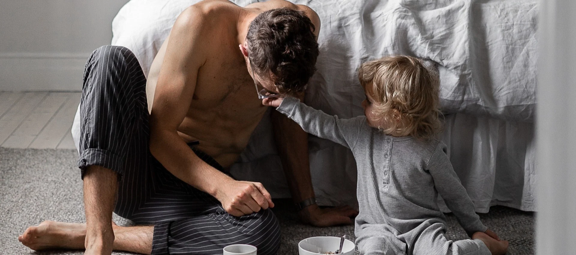 Padre e hijo sentados en el suelo del dormitorio, el niño tocando juguetonamente las gafas del padre, con el desayuno cerca.