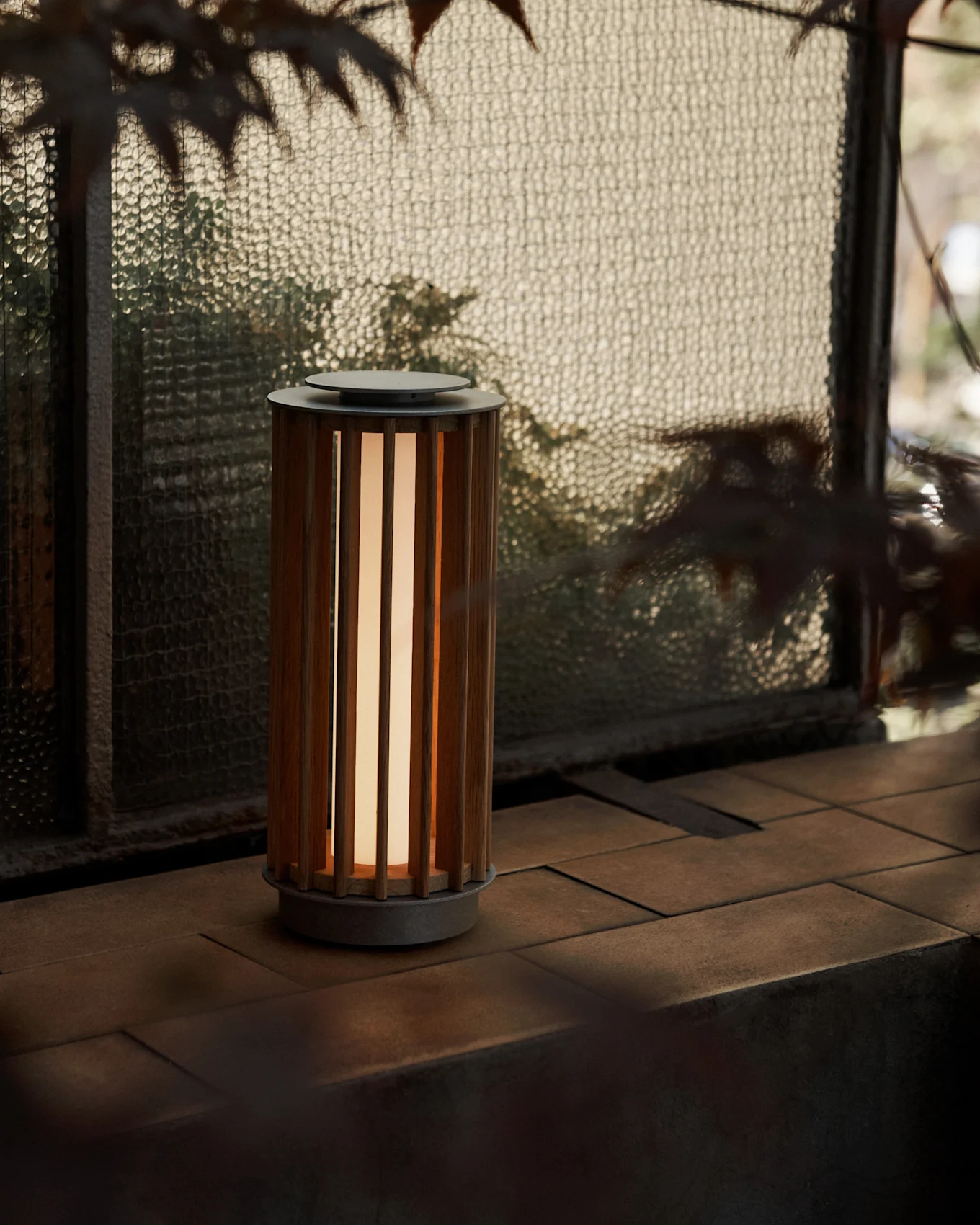 Una lámpara portátil cilíndrica con cuerpo de madera de listones y luz cálida, sobre una superficie de baldosas junto a una ventana de cristal texturizado.