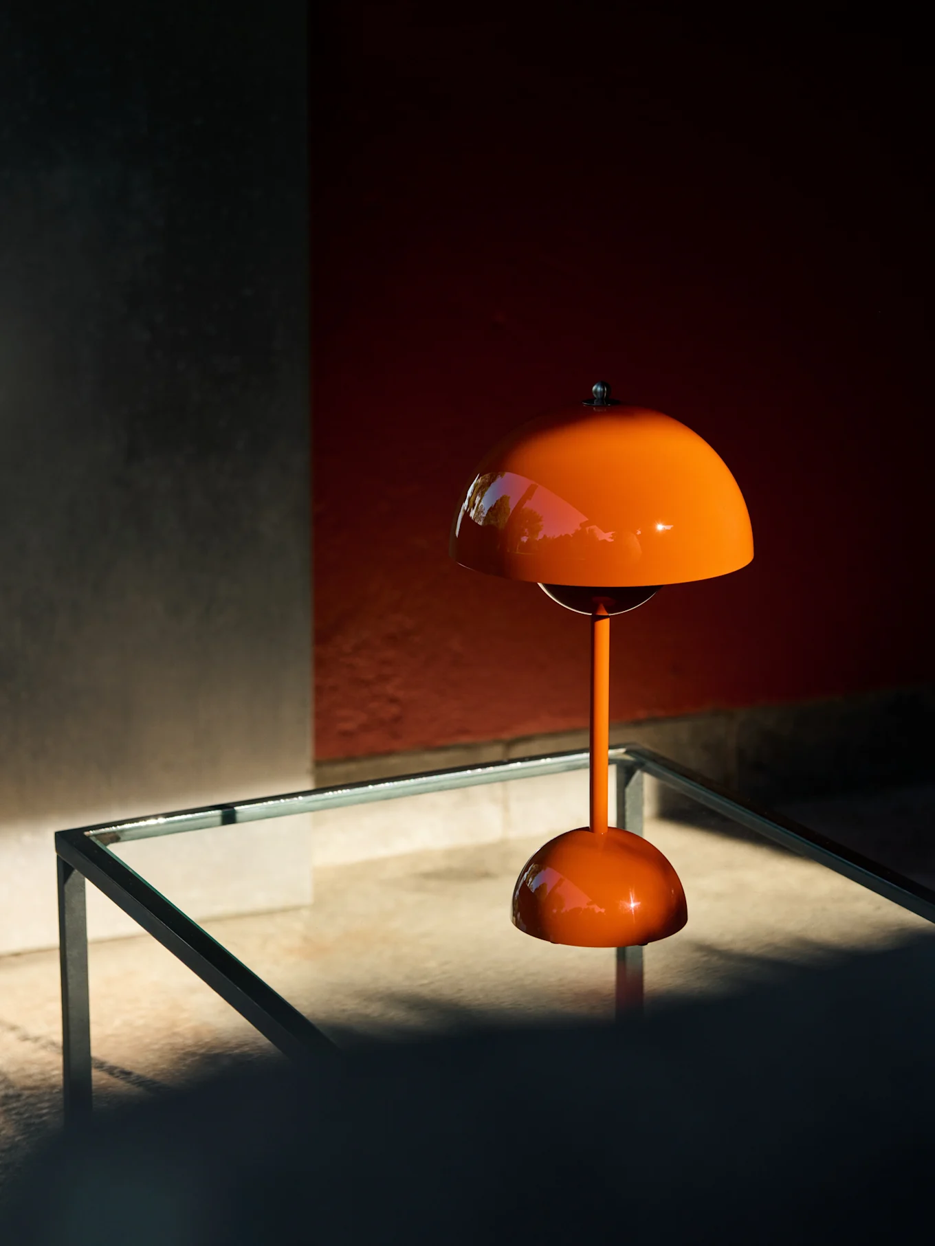 Lámpara de mesa naranja en forma de seta sobre una mesa de cristal, reflejando vegetación.