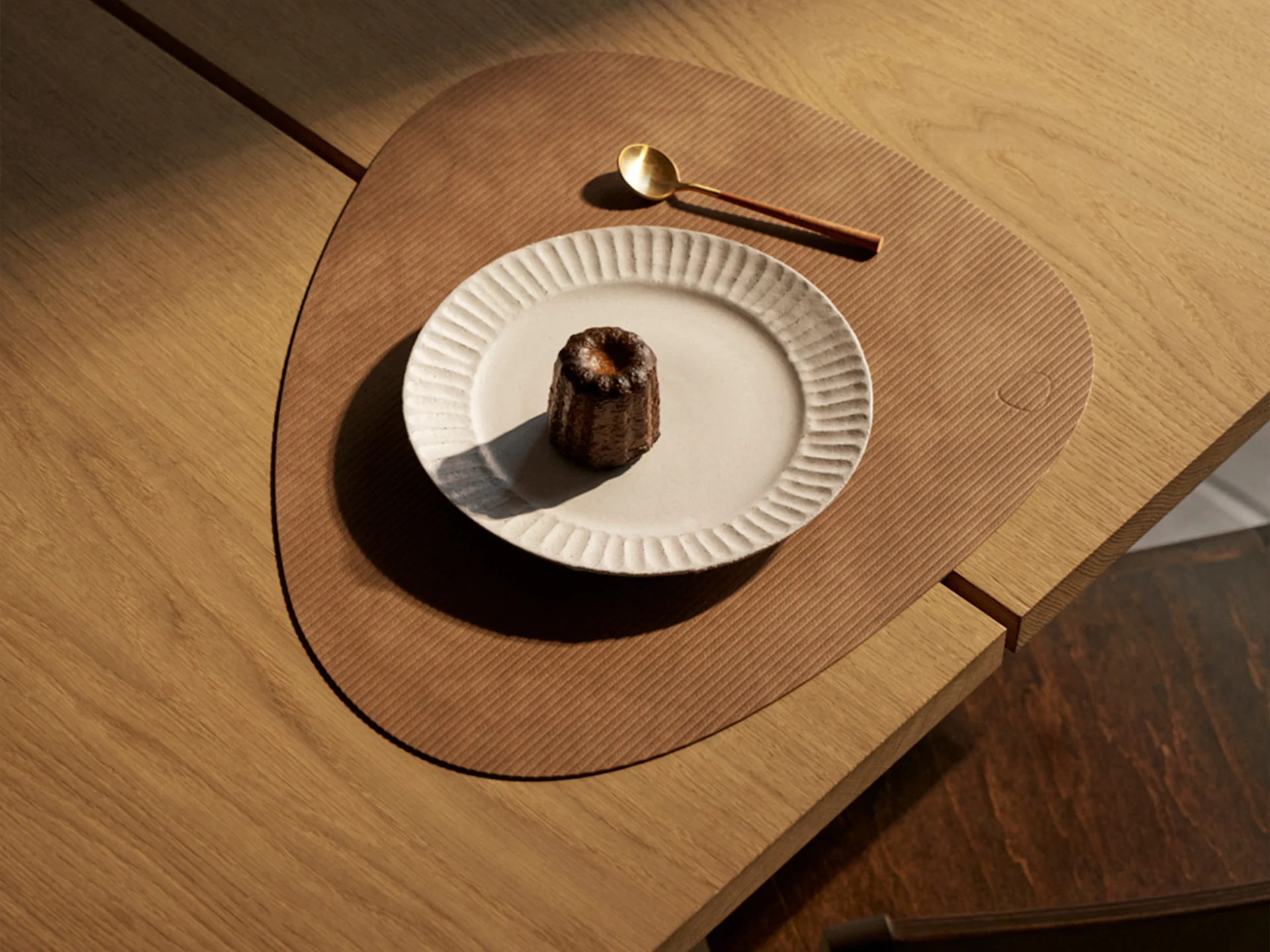 Un canelé sobre un plato acanalado, una cuchara dorada y un salvamanteles triangular en una mesa de madera, iluminado por el sol.