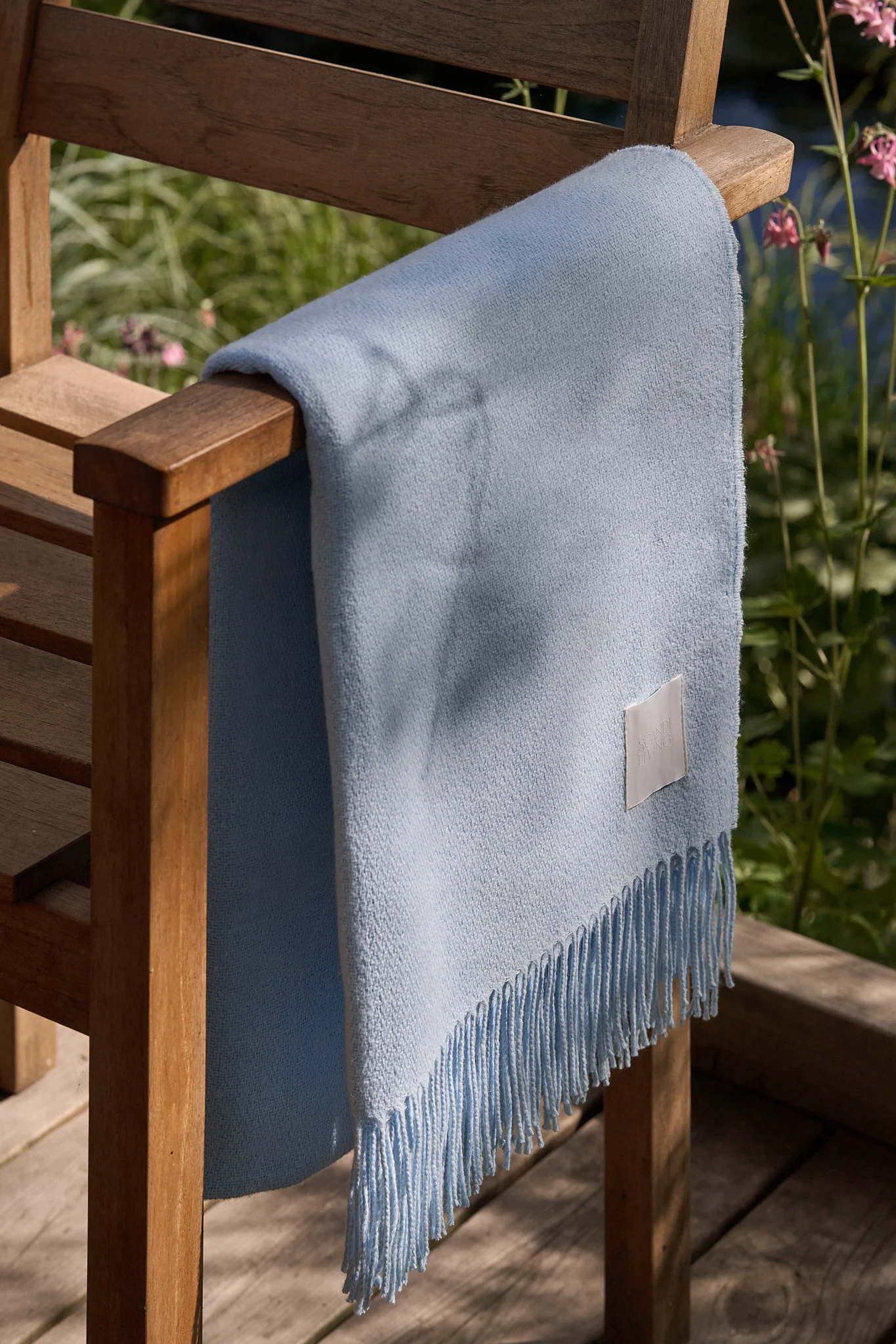 Manta azul claro con flecos sobre una silla de madera de exterior en un jardín soleado.