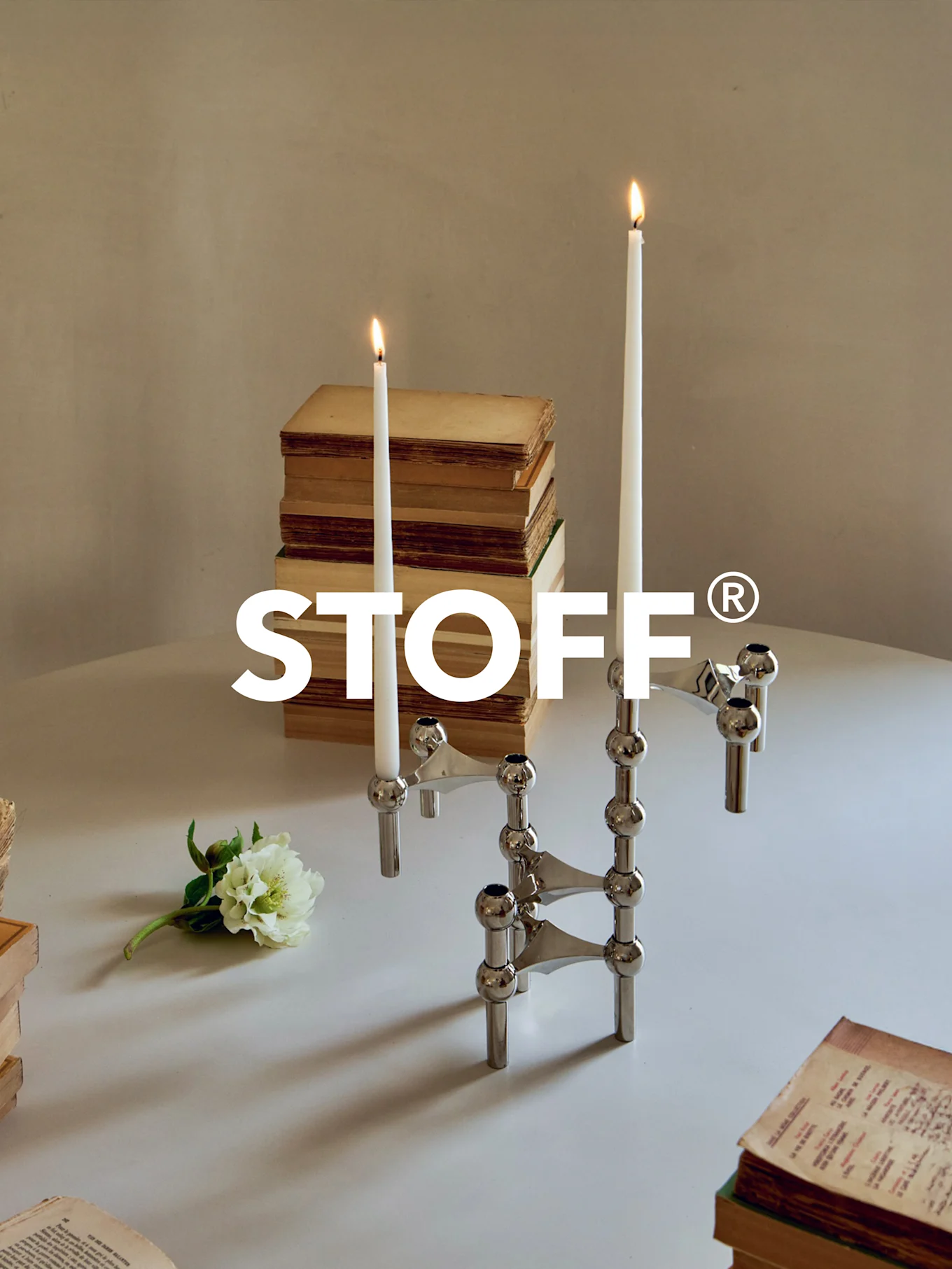 Candelabro modular de cromo pulido con dos velas encendidas, rodeado de libros antiguos y una flor blanca sobre una mesa. Logo STOFF®.