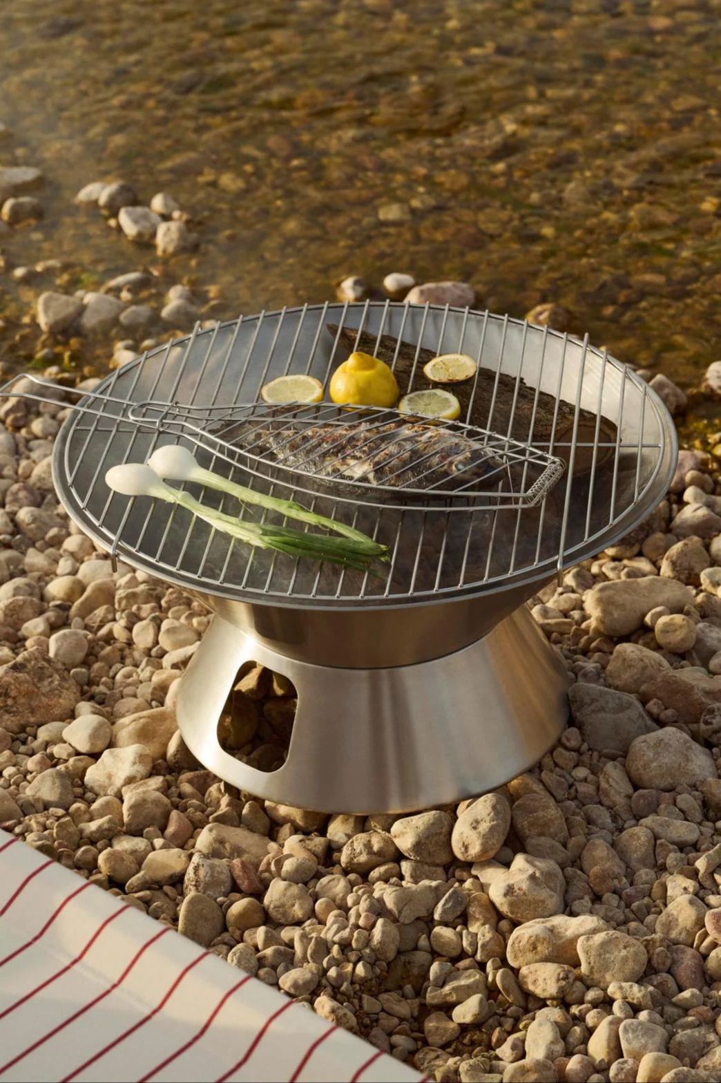 Una parrilla portátil de acero inoxidable con un pescado entero en una cesta para asar, limones y cebolletas en una orilla rocosa.