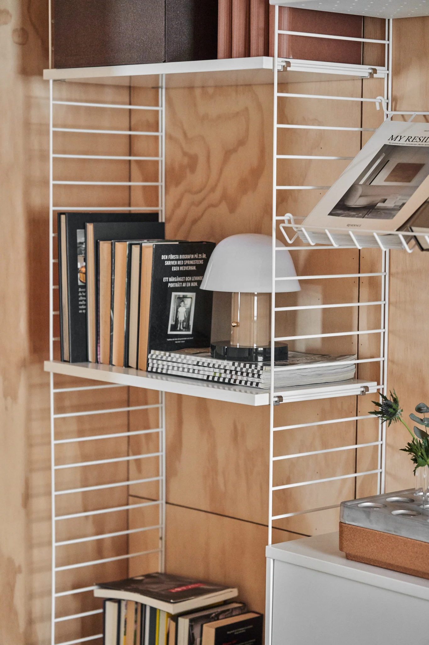 Estantería de alambre blanco en una pared de madera, con libros, una lámpara de mesa blanca y una revista. Un mueble gris está debajo.