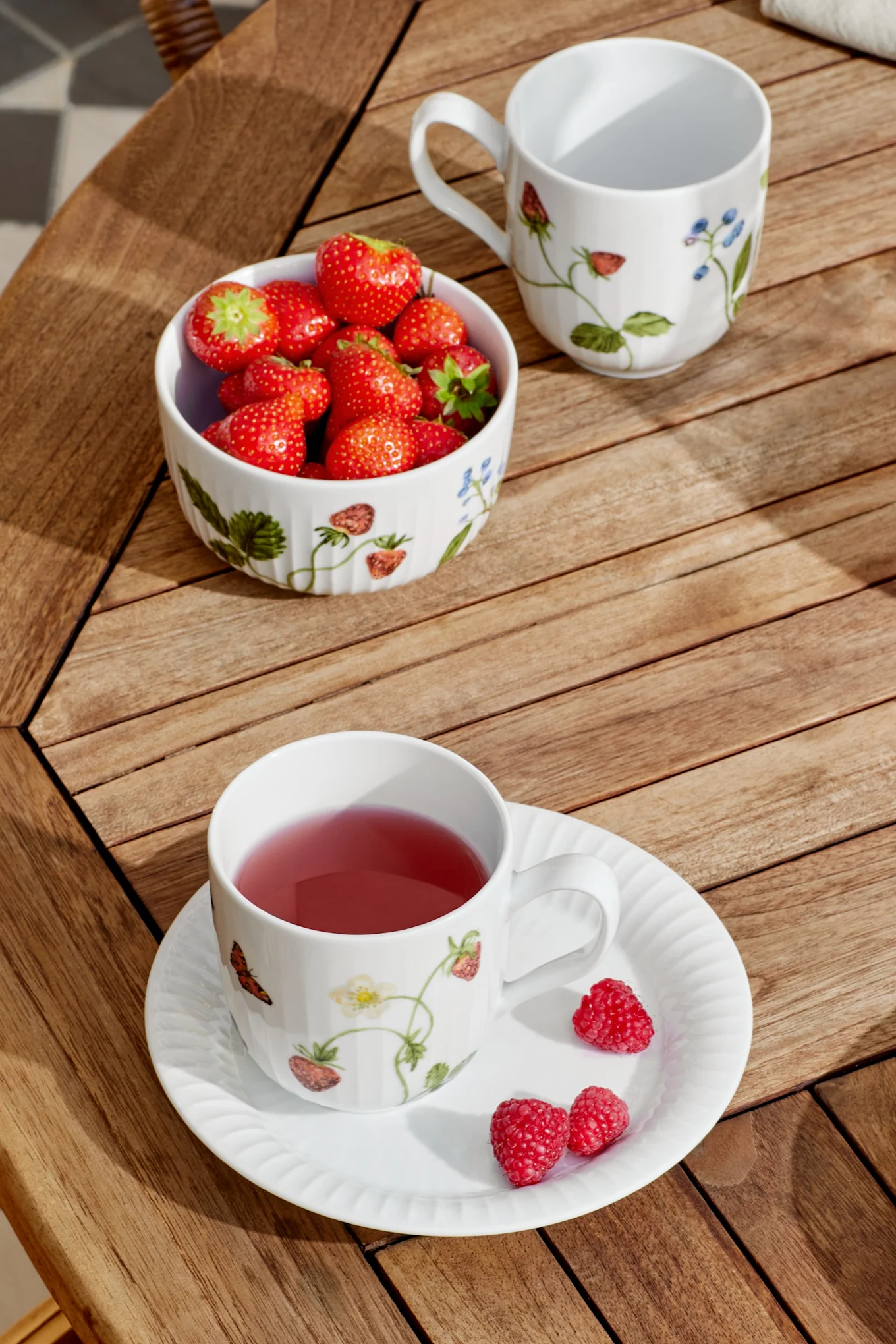 Una taza blanca con té rosa y un platillo con frambuesas, un tazón de fresas y una taza vacía sobre una mesa de madera en la luz del sol.