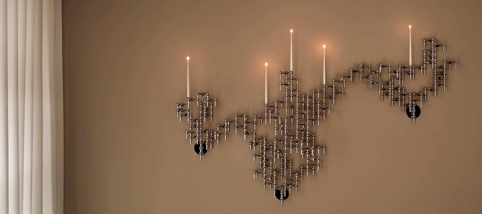 Un candelabro modular plateado montado en la pared con varias velas encendidas sobre una pared beige, junto a persianas verticales.
