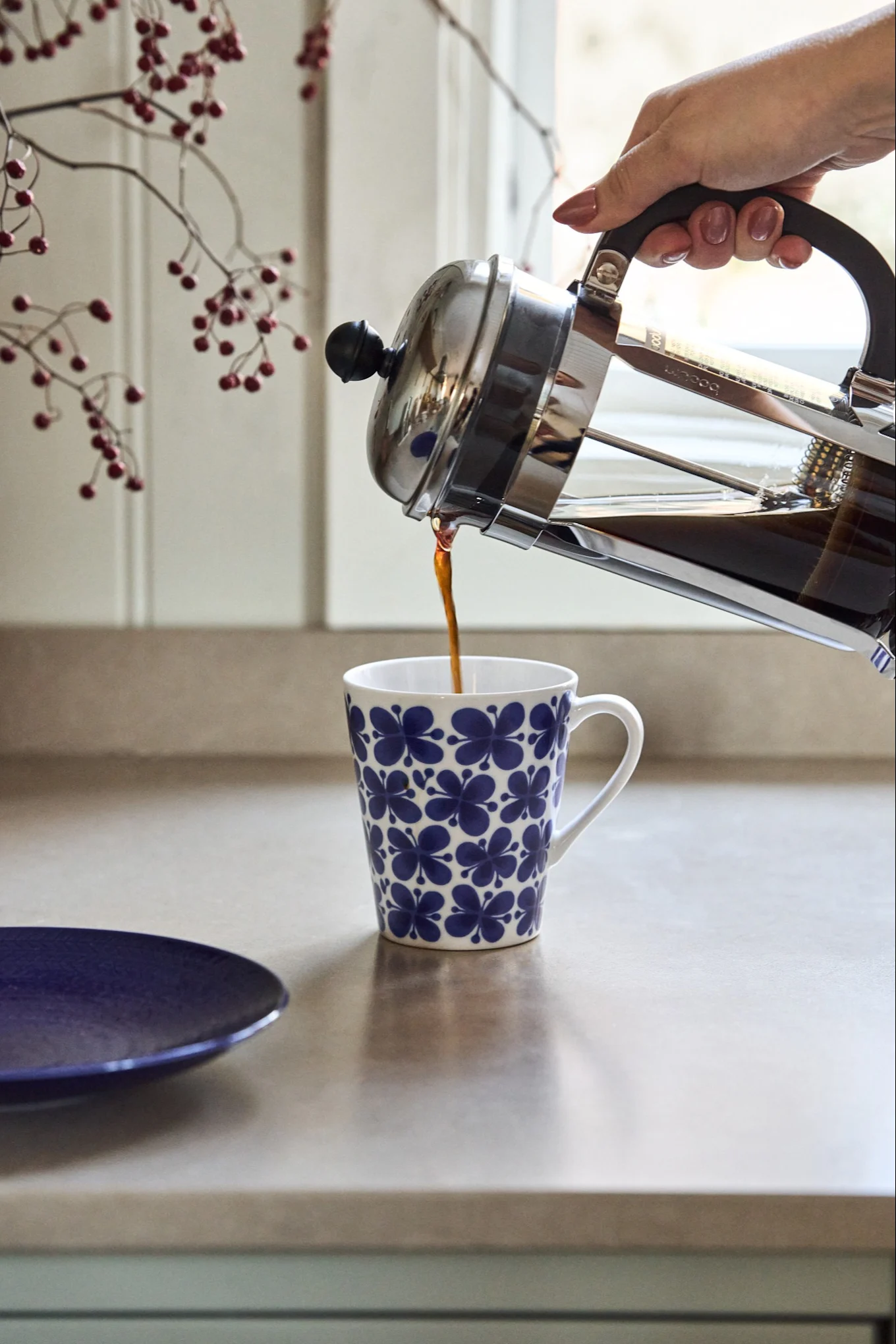 Una mano vierte café de una cafetera francesa en una taza blanca con un patrón floral azul sobre una encimera de cocina.