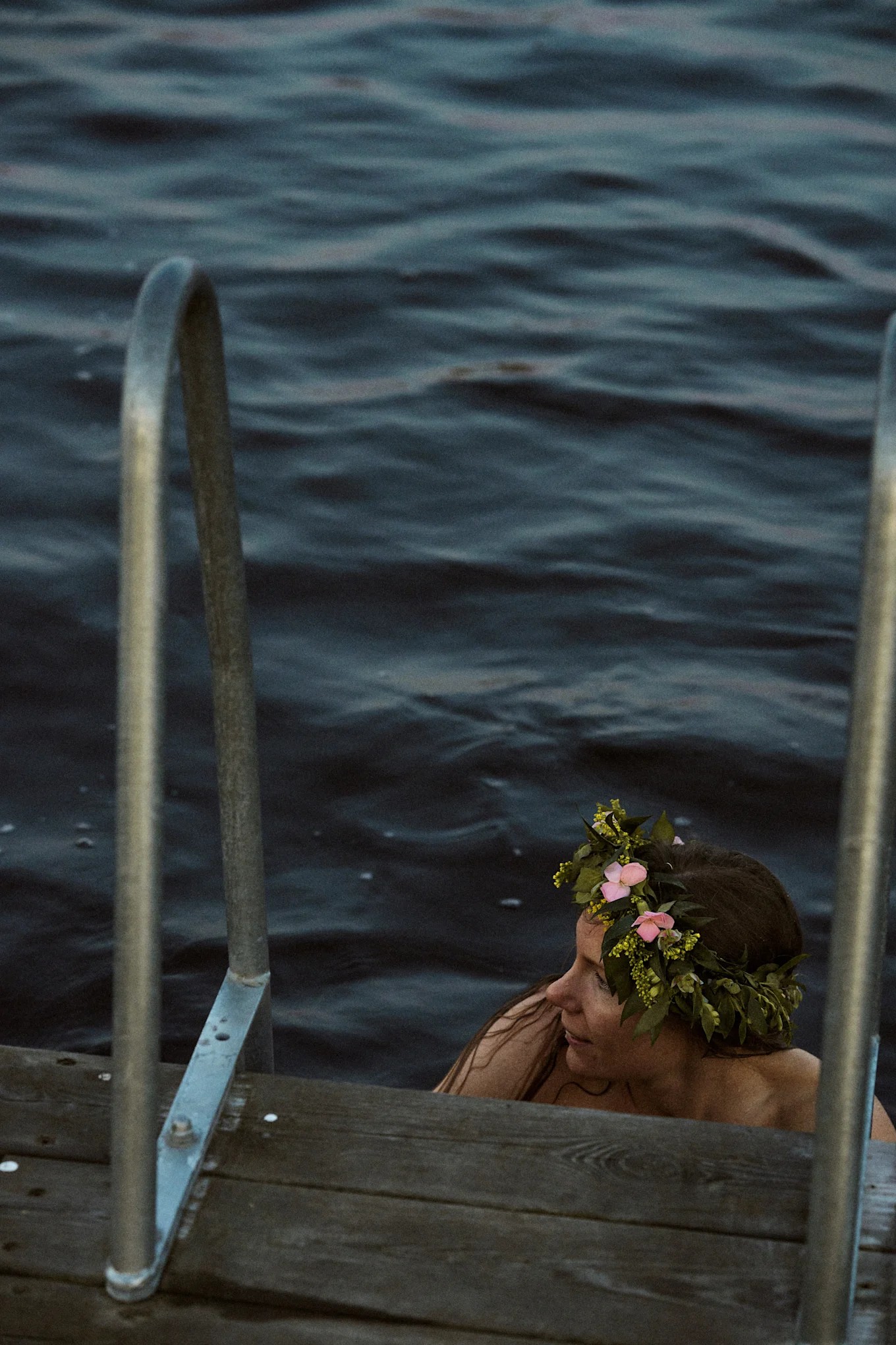 Mujer en agua oscura con corona de flores, junto a un muelle de madera con pasamanos de metal.