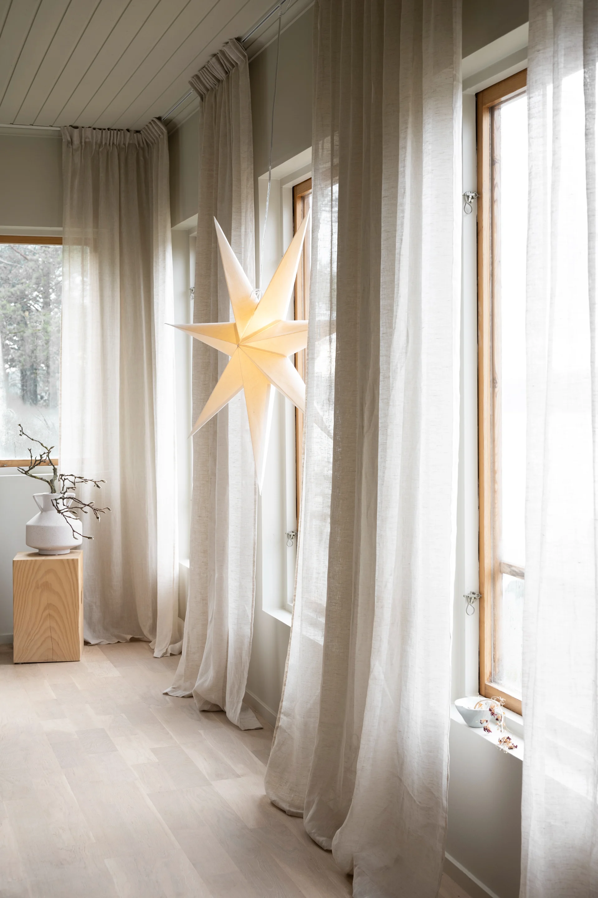 Una estrella de papel iluminada cuelga en una ventana con cortinas transparentes, junto a un jarrón blanco con ramas sobre un bloque de madera.