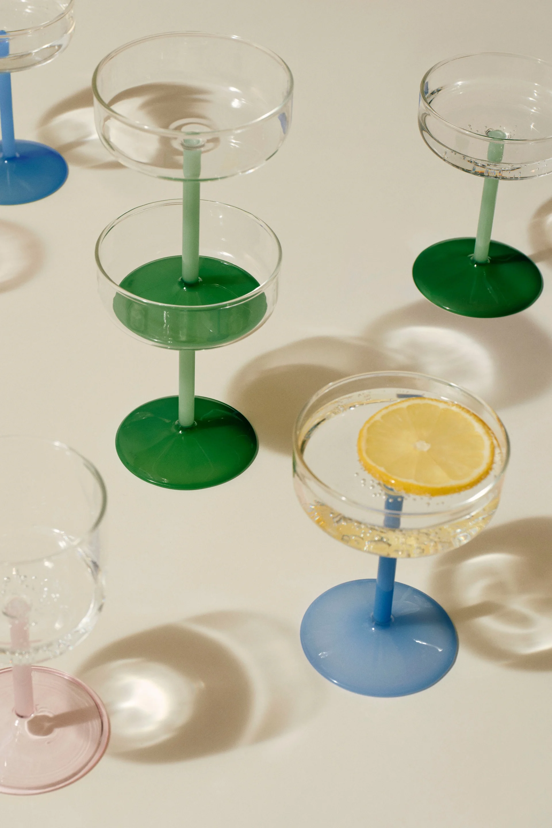 Surtido de copas coupé con tallos y bases de colores. Una copa contiene una rodaja de limón en agua con gas.