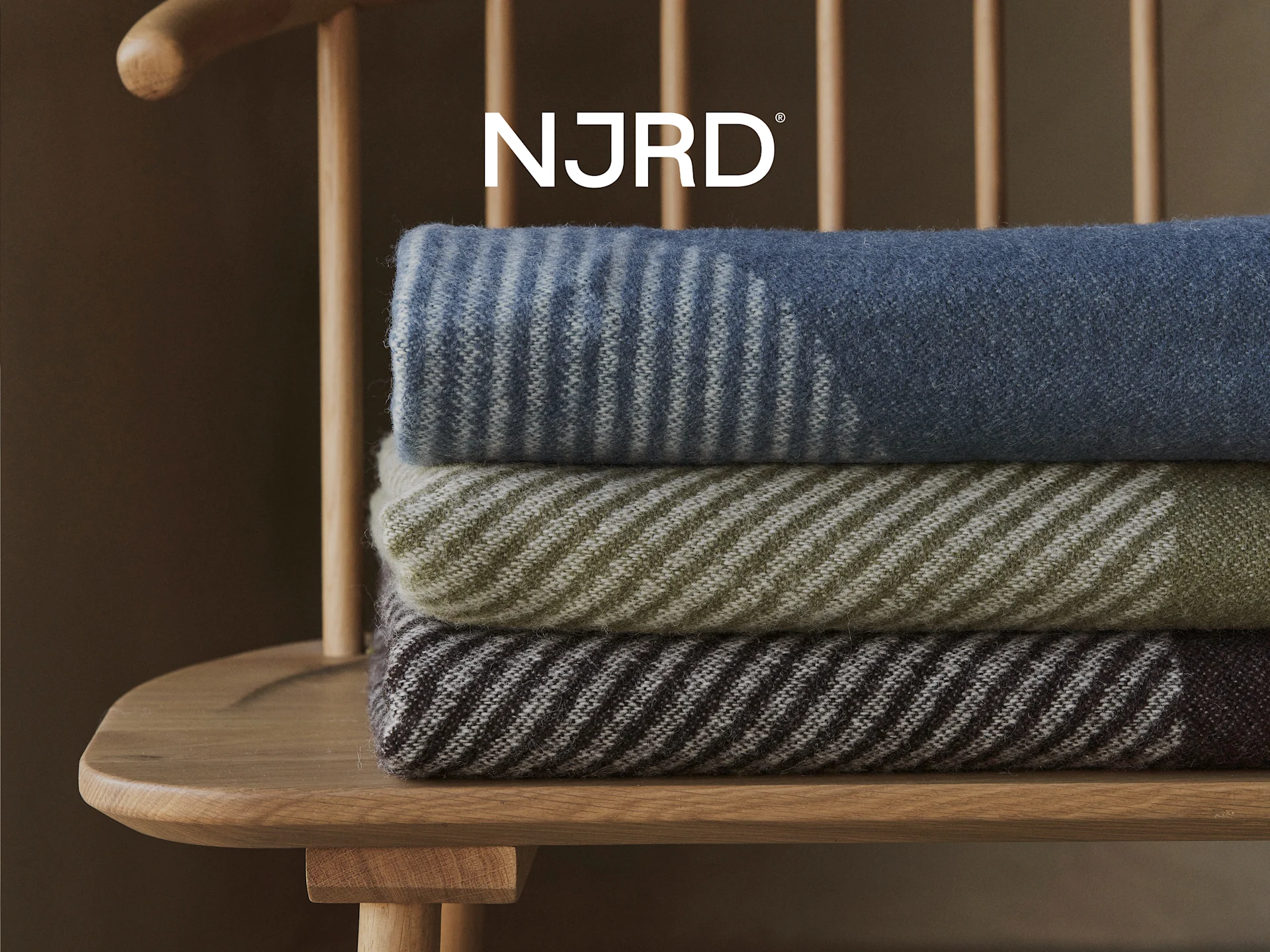 Tres mantas a rayas en azul, oliva y marrón, apiladas en una silla de madera con el logo de NJRD encima.