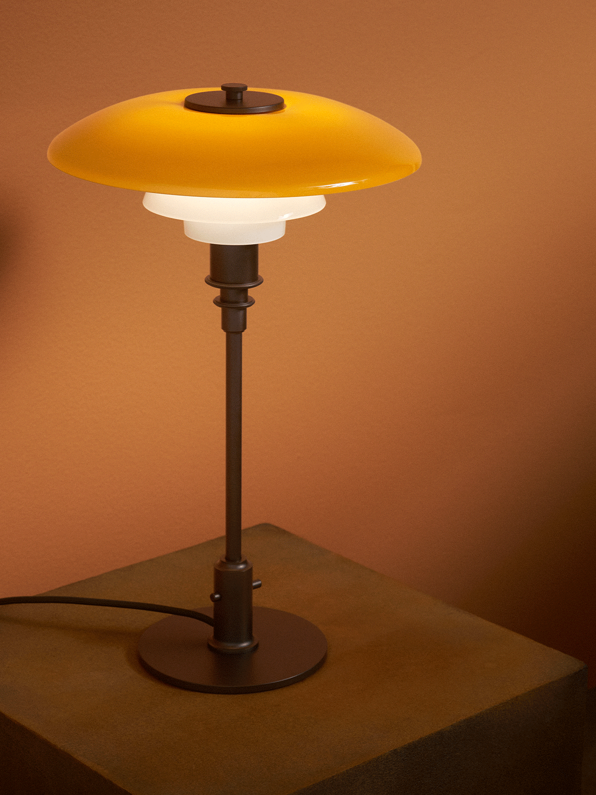 Lámpara de mesa moderna con pantalla superior amarilla y dos blancas, sobre un pedestal de madera texturizado, contra pared marrón.