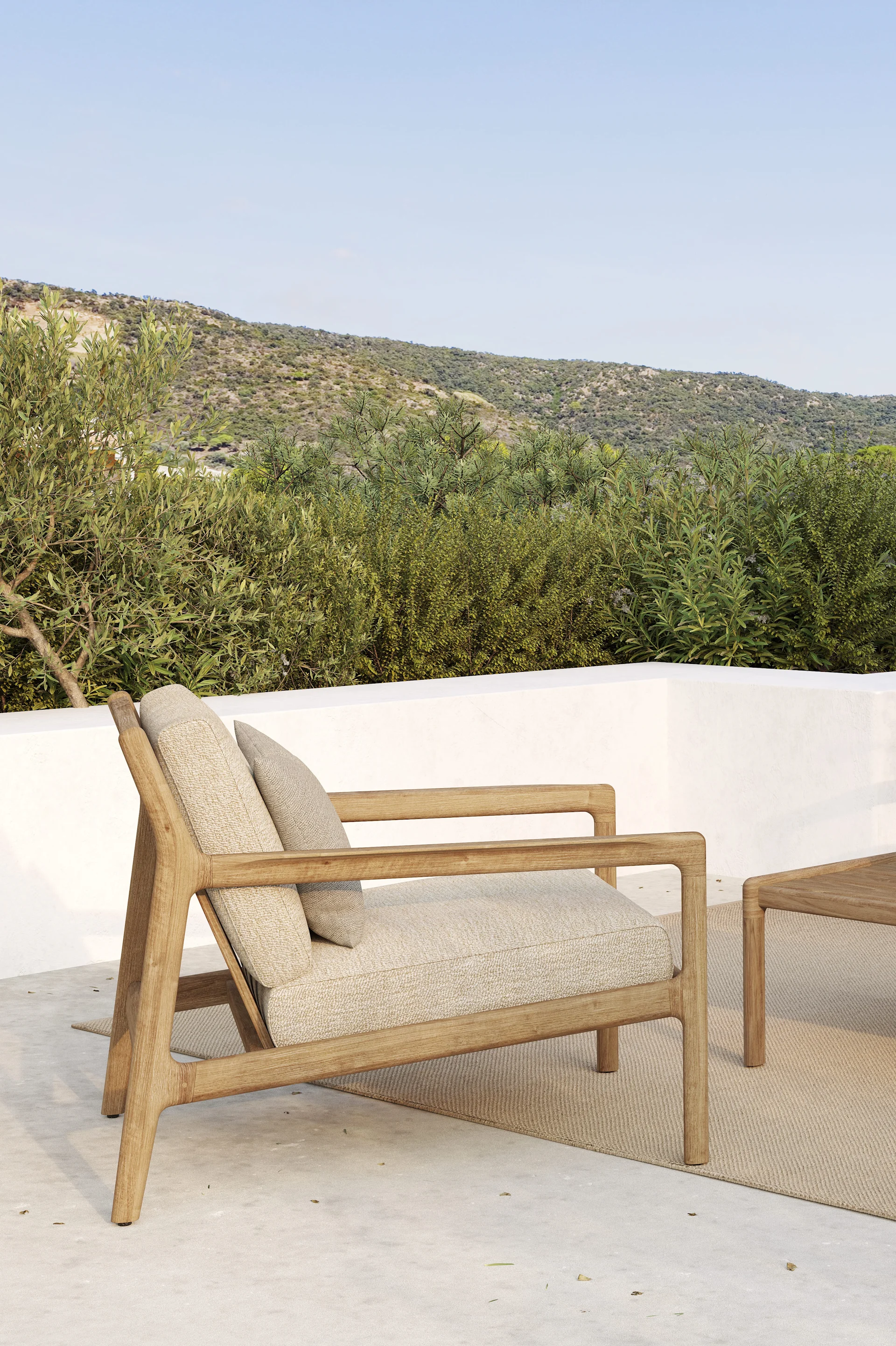 Sillón de exterior de madera con cojines claros en un patio, con colinas verdes y cielo azul de fondo.