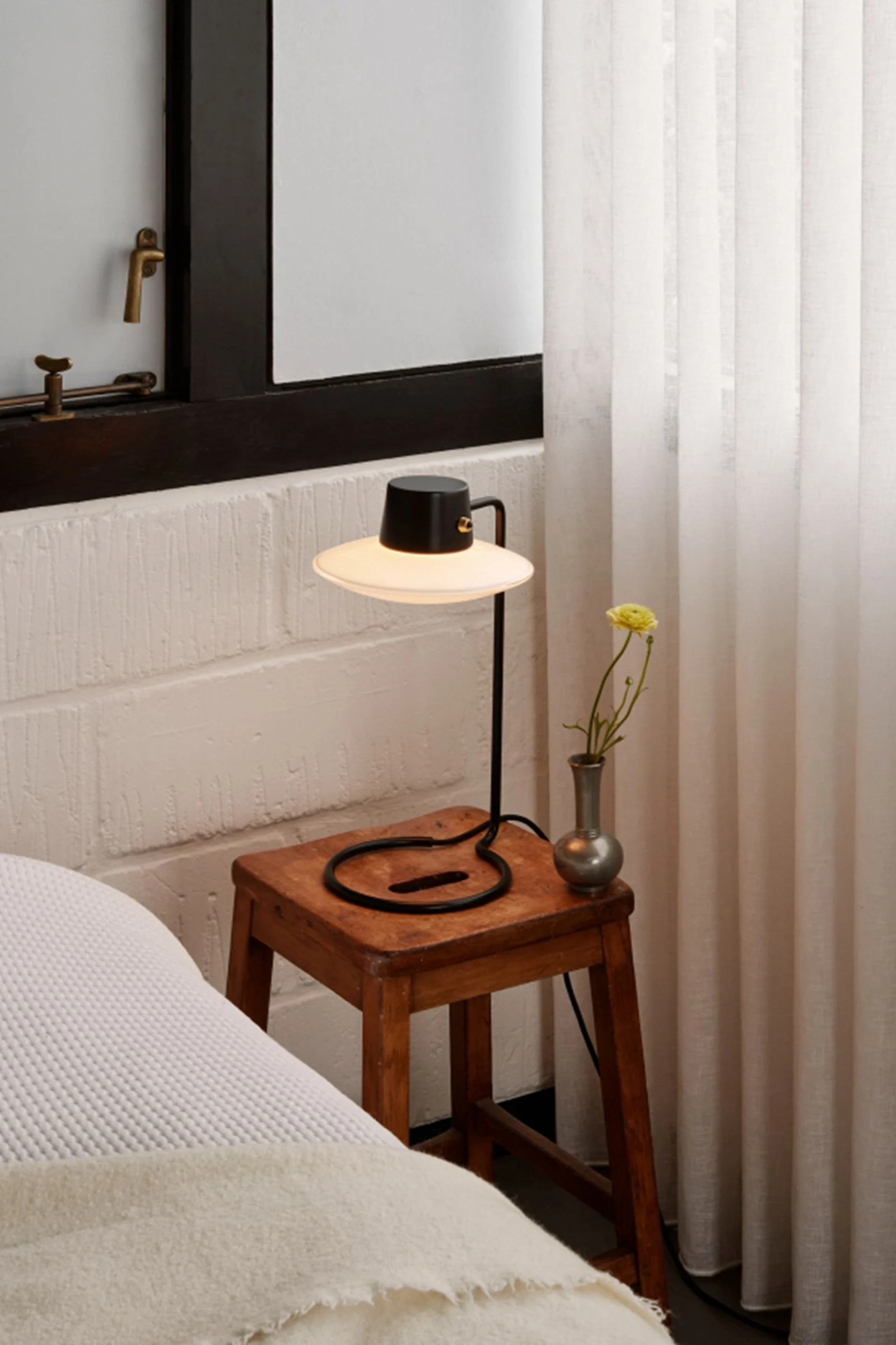 Lámpara de mesa negra con pantalla ancha en taburete rústico de madera, jarrón con flor amarilla, junto a cama y cortinas.